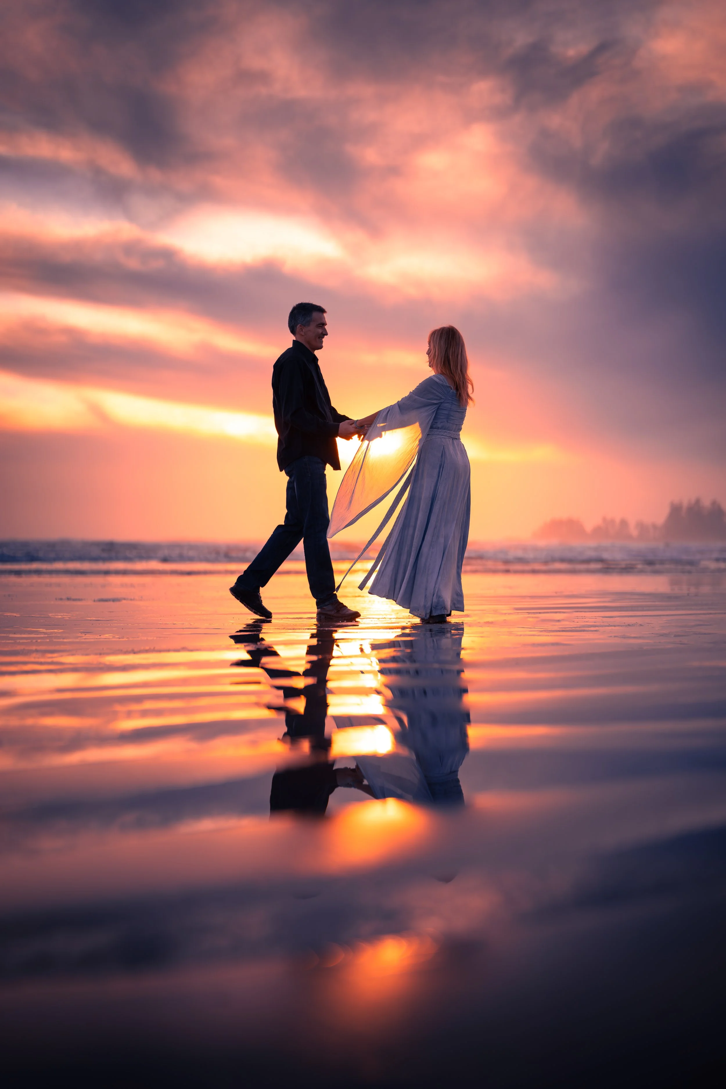 A couple holds hands on a beach at sunset, with reflections on the water.