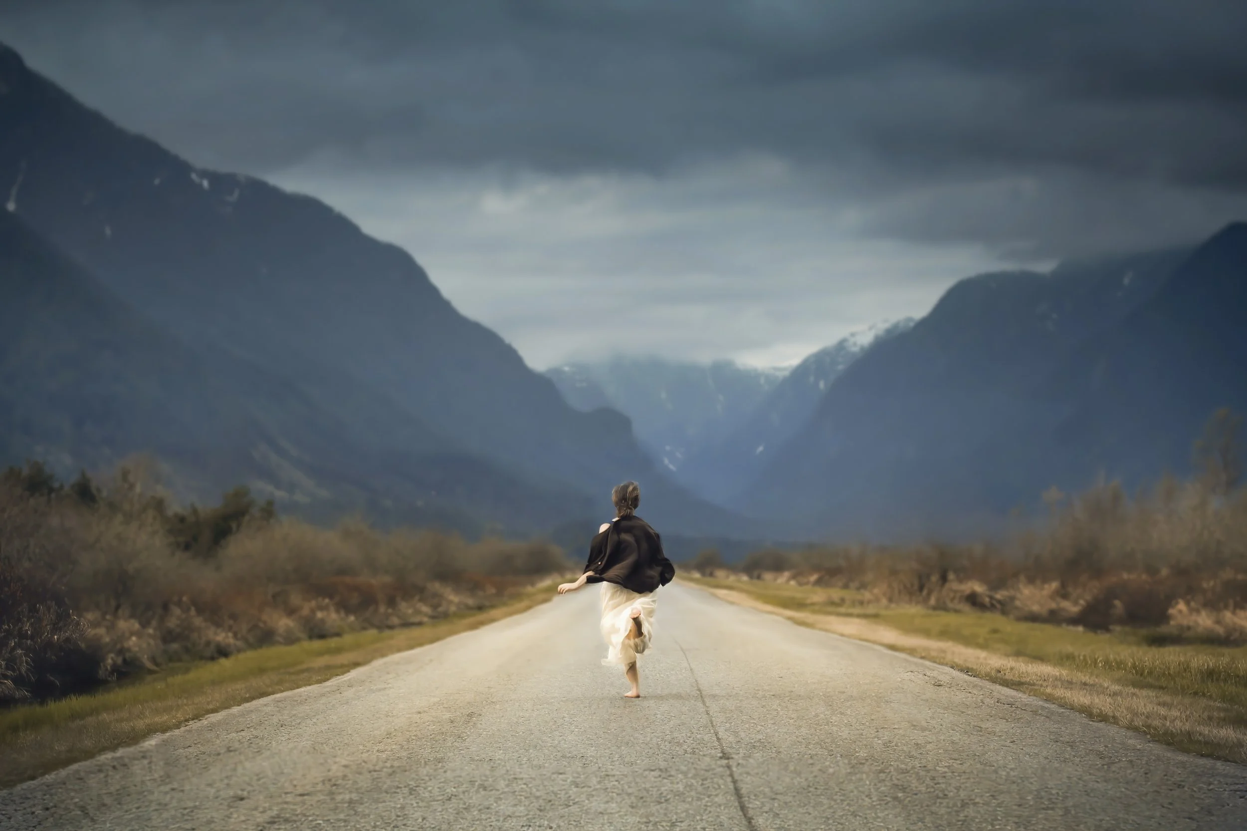 Person walking barefoot on a deserted road surrounded by mountains and cloudy sky.