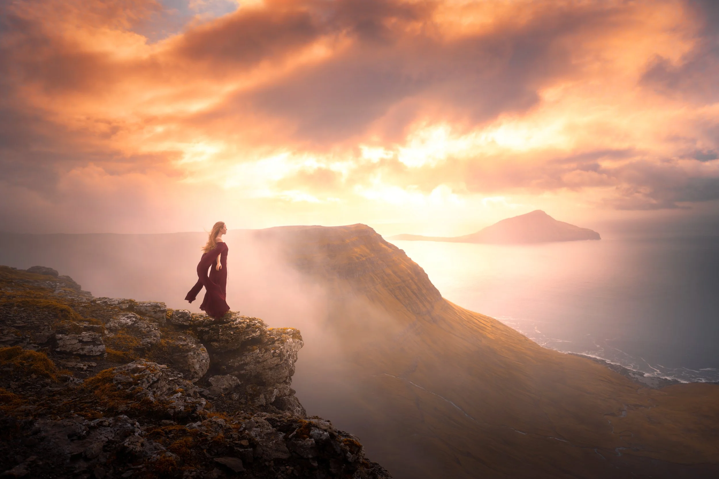 A woman in a dark dress standing on the edge of a rocky cliff at sunrise or sunset, overlooking a misty landscape with hills, water, and islands in the distance.