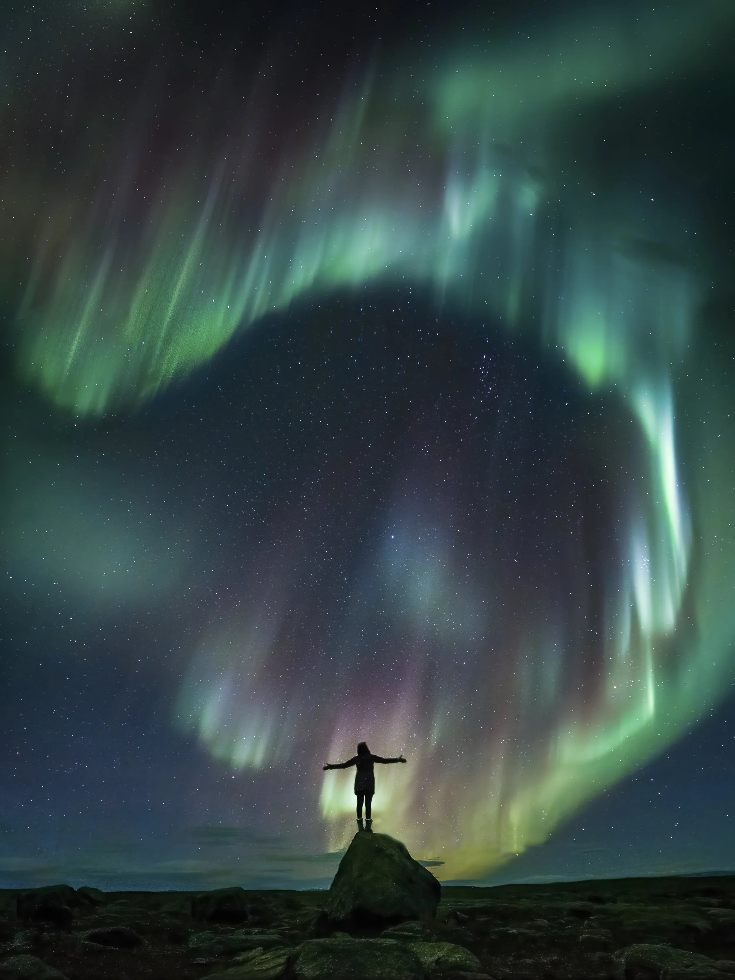 Person standing on a rock with arms outstretched under the Northern Lights and starry night sky.