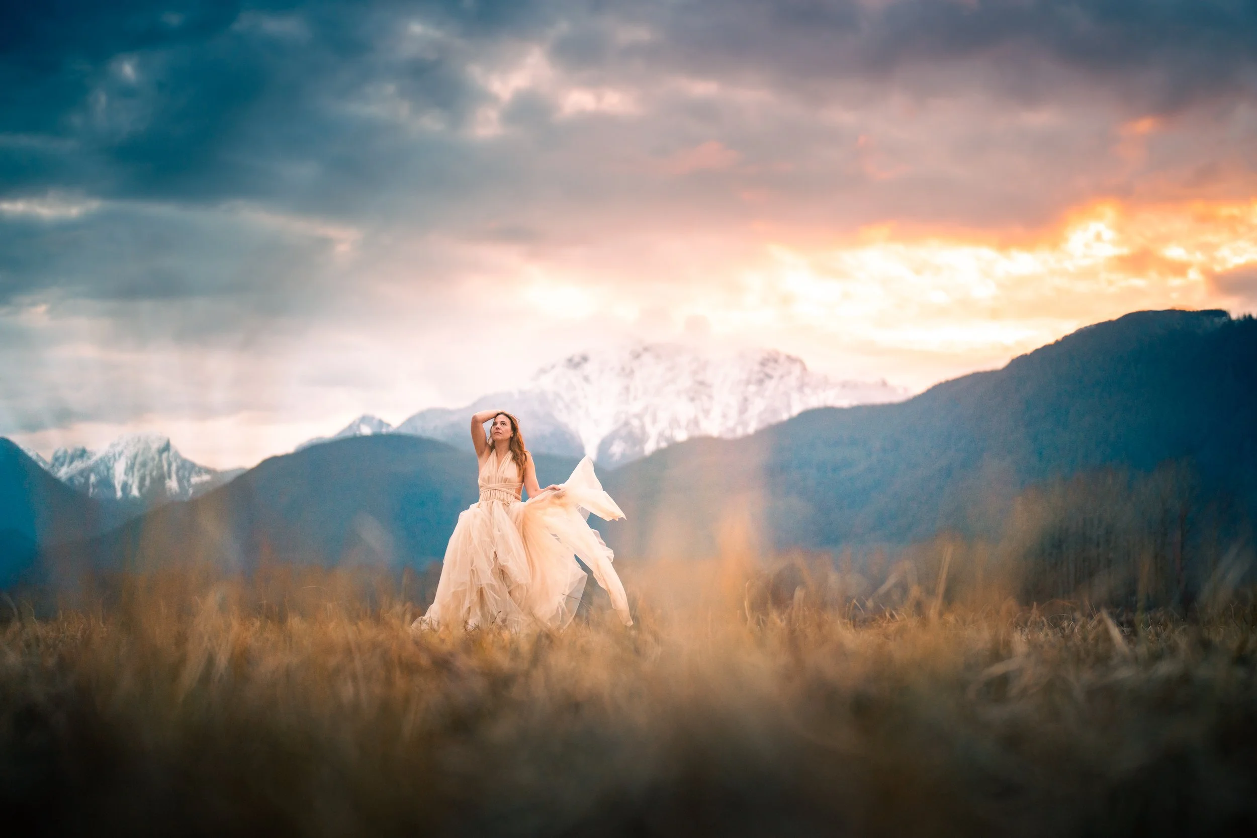 A woman in a flowing beige dress standing in a field with mountains and a colorful sunset sky in the background.