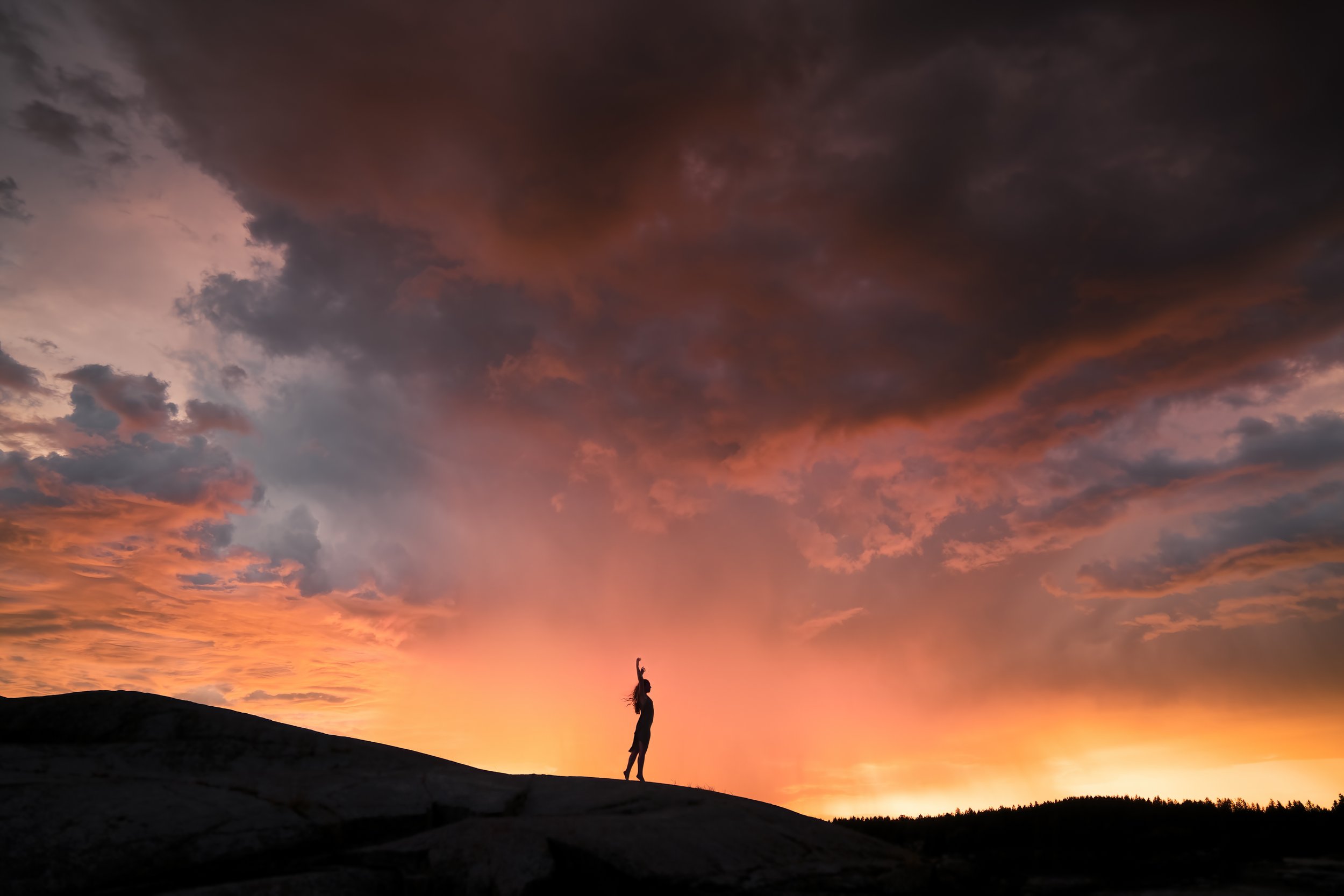 Person standing on a hill during sunset with a sky full of dark and colorful clouds.