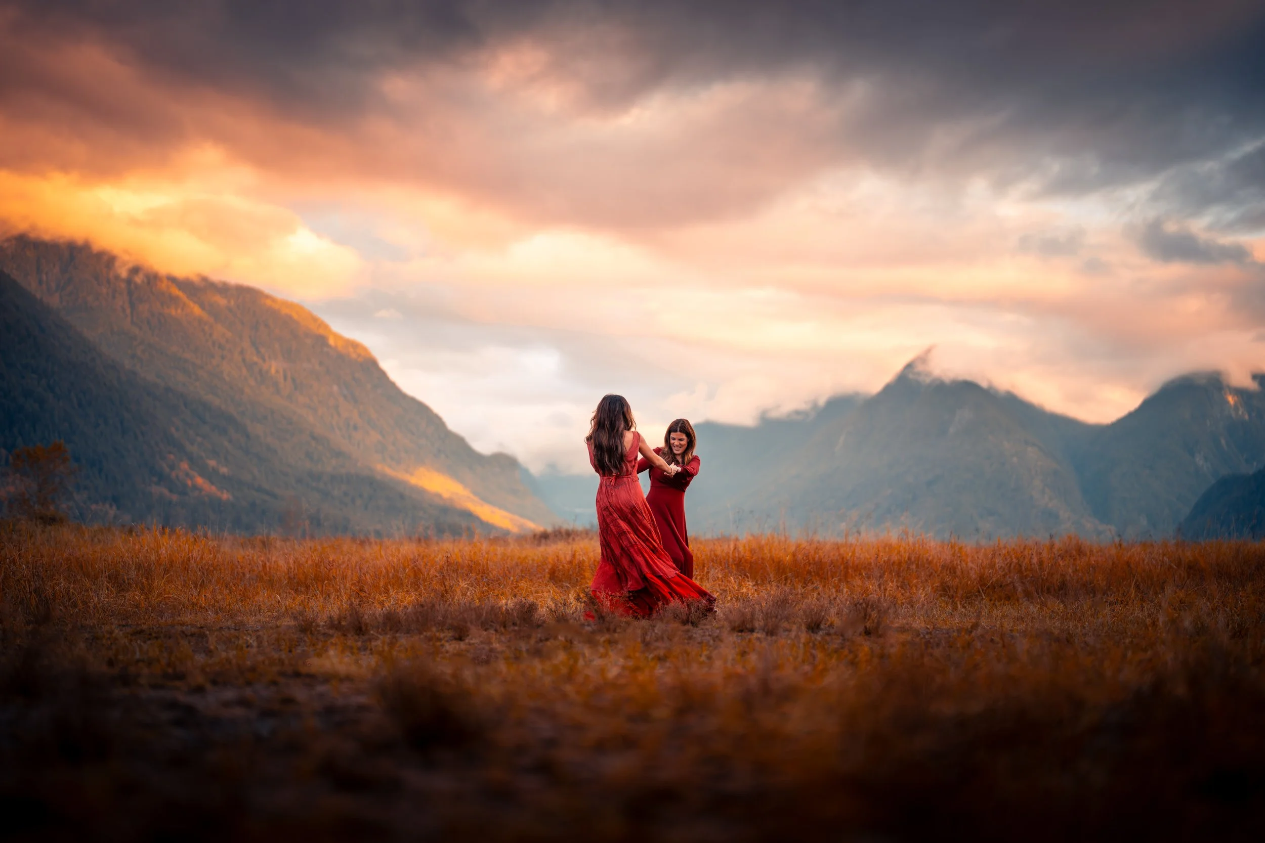 Two women in red dresses dancing in a field at sunset or sunrise with mountains in the background.