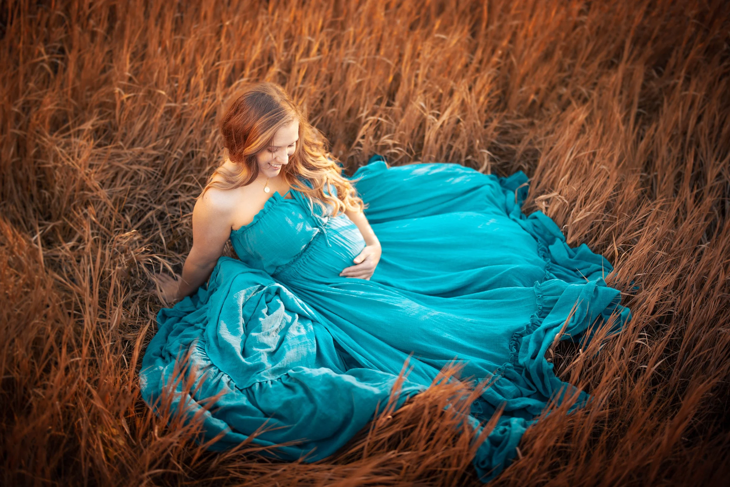 A pregnant woman in a flowing turquoise dress sitting in a field of dry, brown grass, smiling and gently touching her belly.