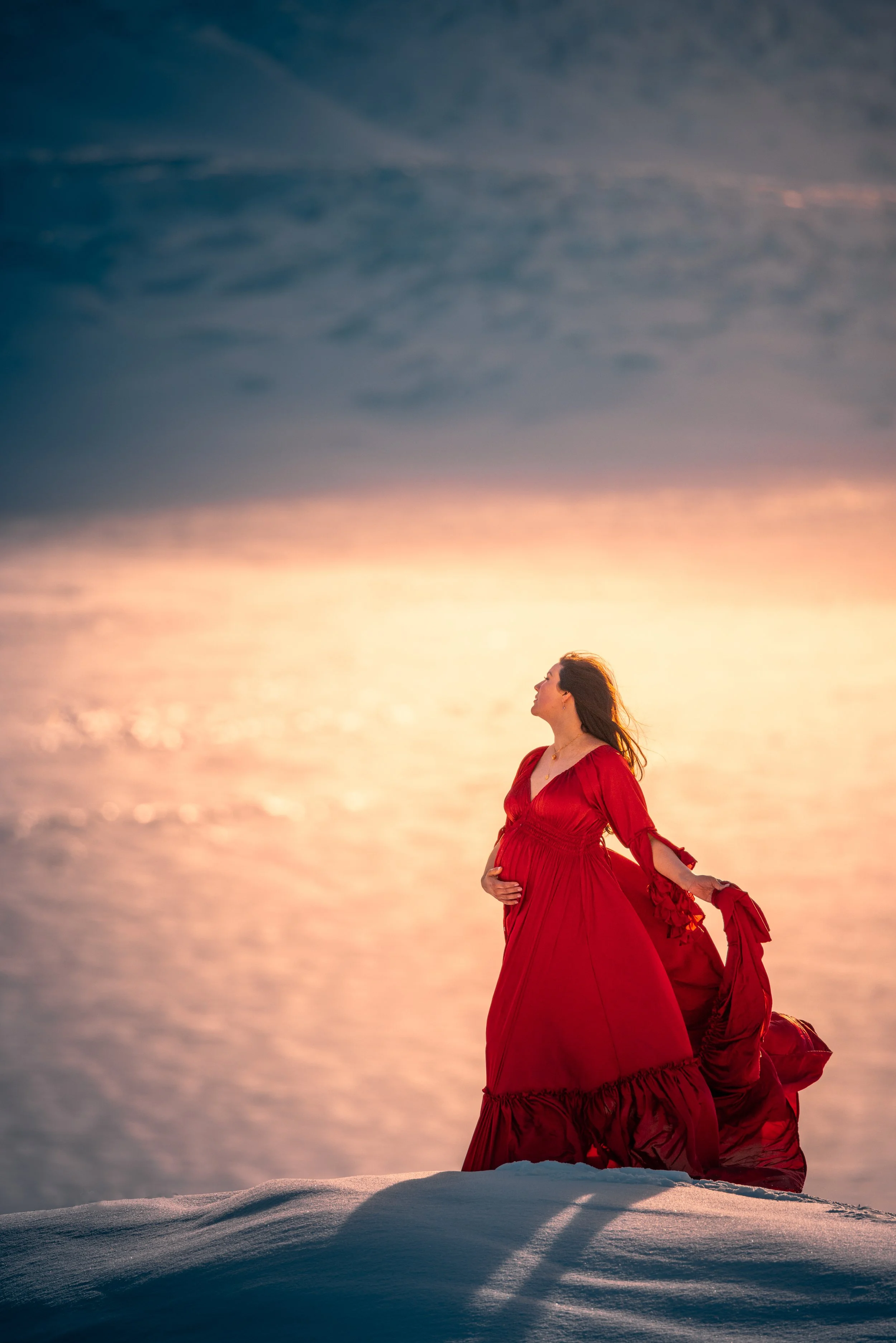 A pregnant woman in a flowing red dress stands on snow-covered ground, looking up as the sunset reflects on a body of water in the background.