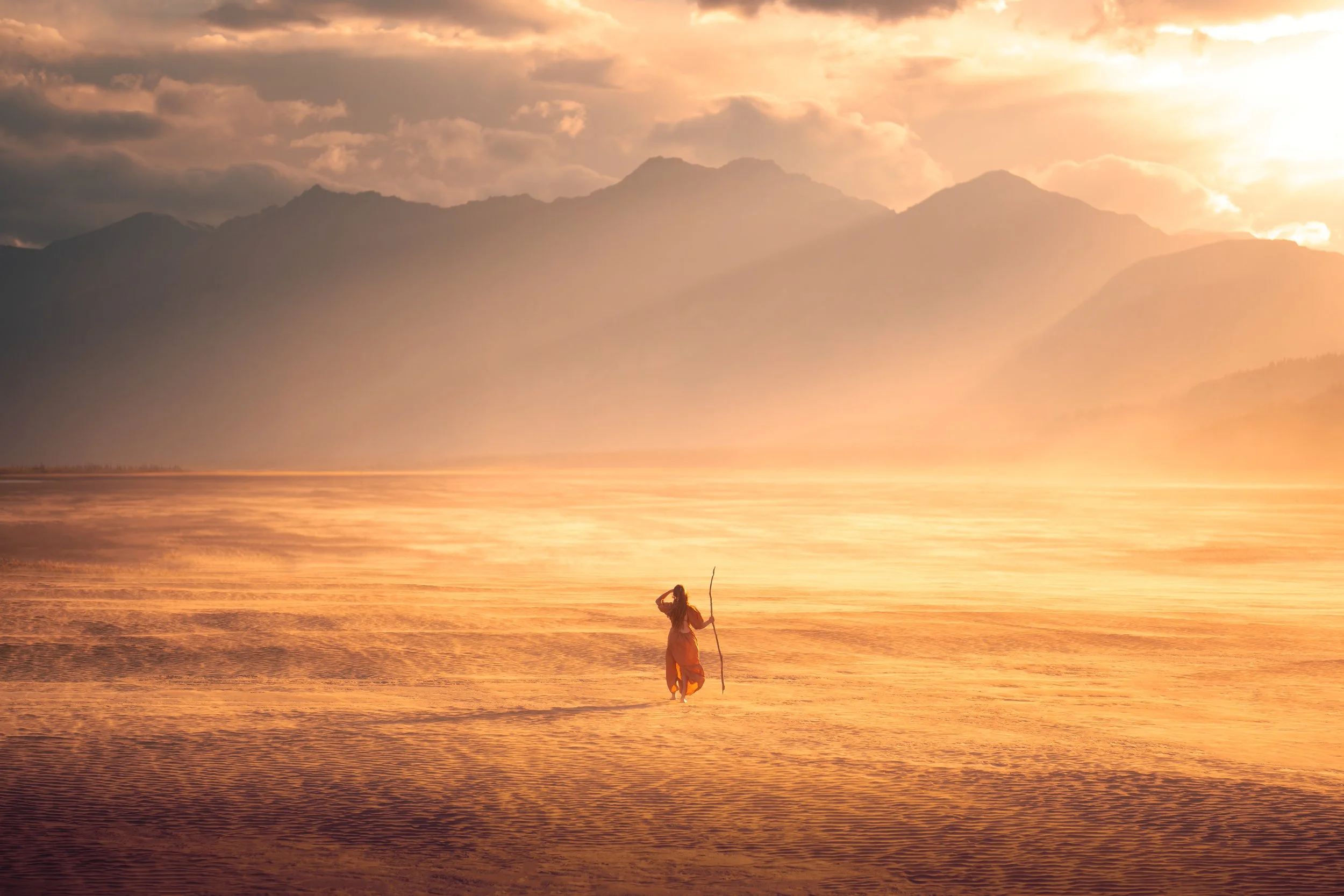 A person walking on a calm body of water holding a stick, with mountains in the background at sunset.