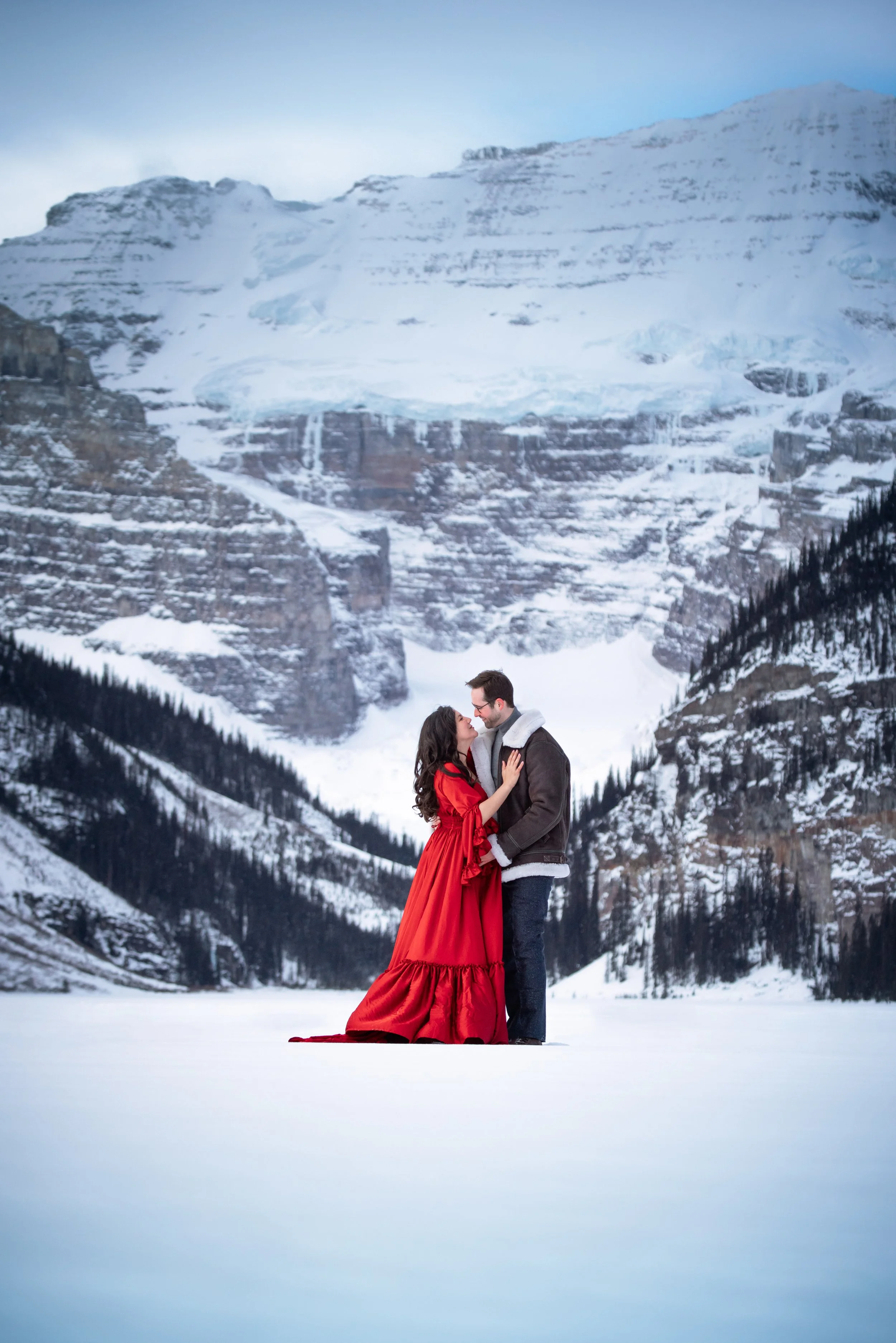 A couple standing close together in a snowy landscape with mountains in the background, the woman wearing a red dress and the man in a dark jacket.