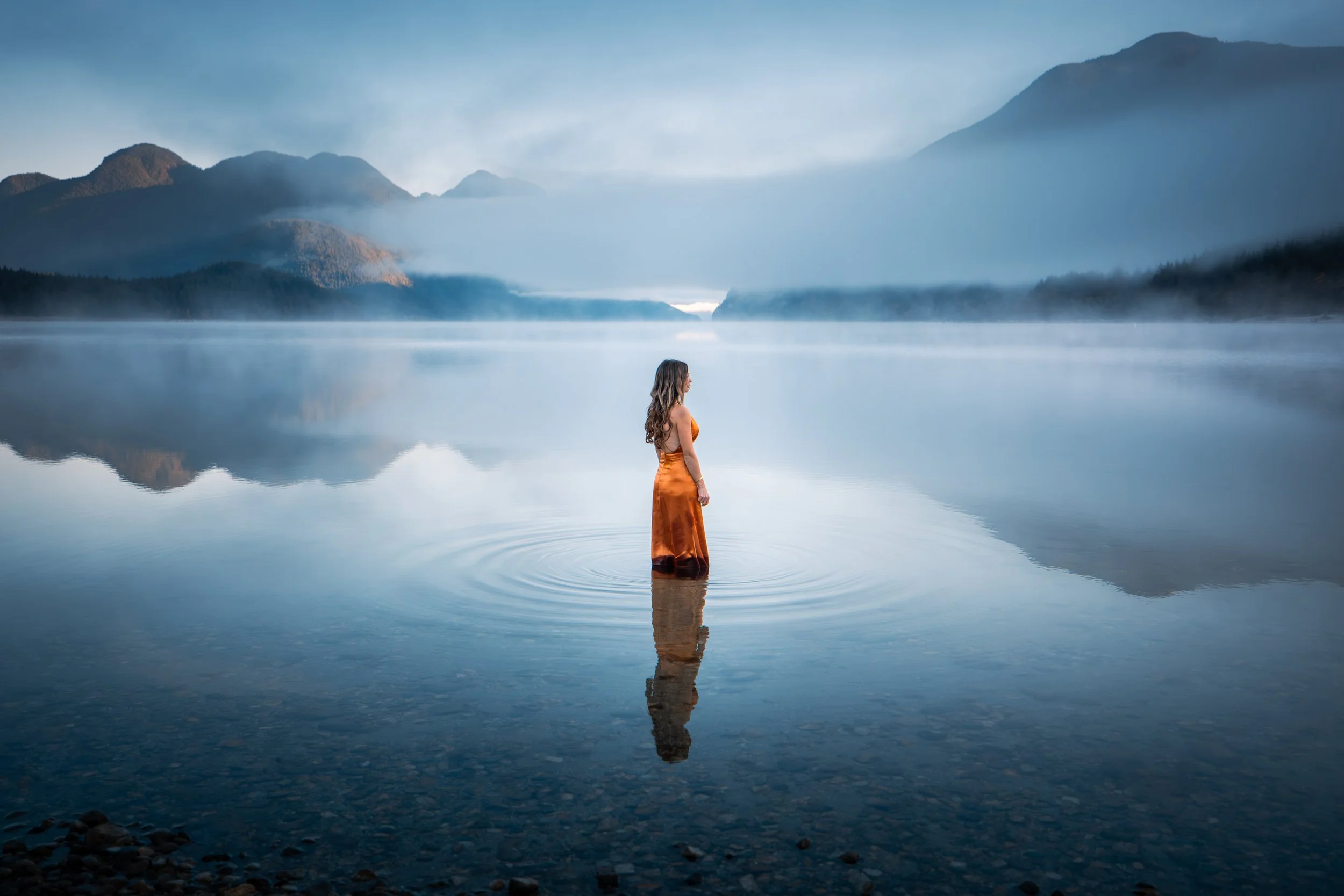A woman standing in a calm lake with mountains in the background and fog, wearing an orange dress.