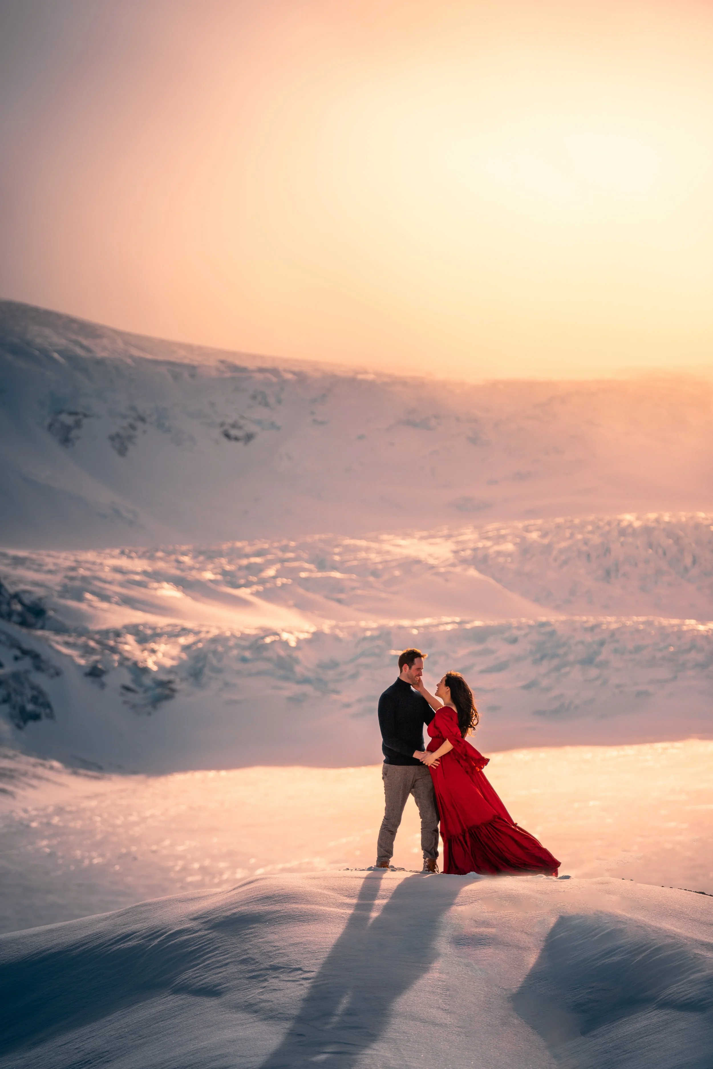 A couple standing on snow-covered ground with a snowy mountain landscape and setting sun in the background. The woman wears a flowing red dress, and the man wears a black jacket and gray pants.