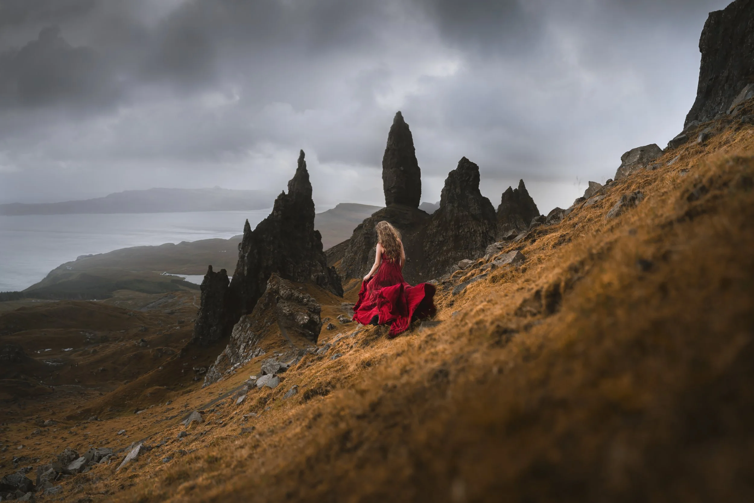 A woman in a flowing red dress sitting on a grassy hillside with rocky formations in the background under a cloudy sky.