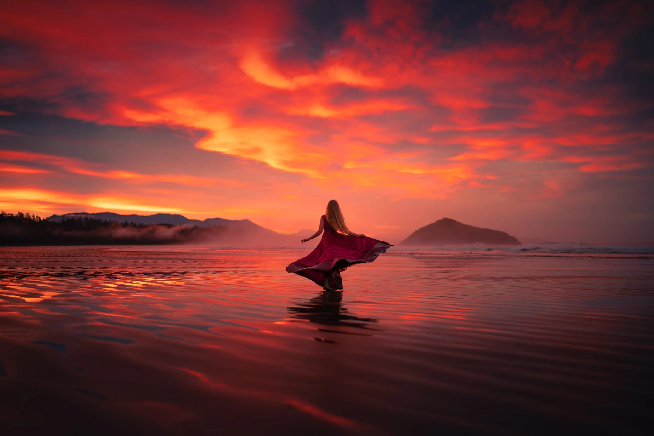 A woman in a flowing red dress wading in the shallow water on a beach during sunset with an orange and pink sky and distant mountains.