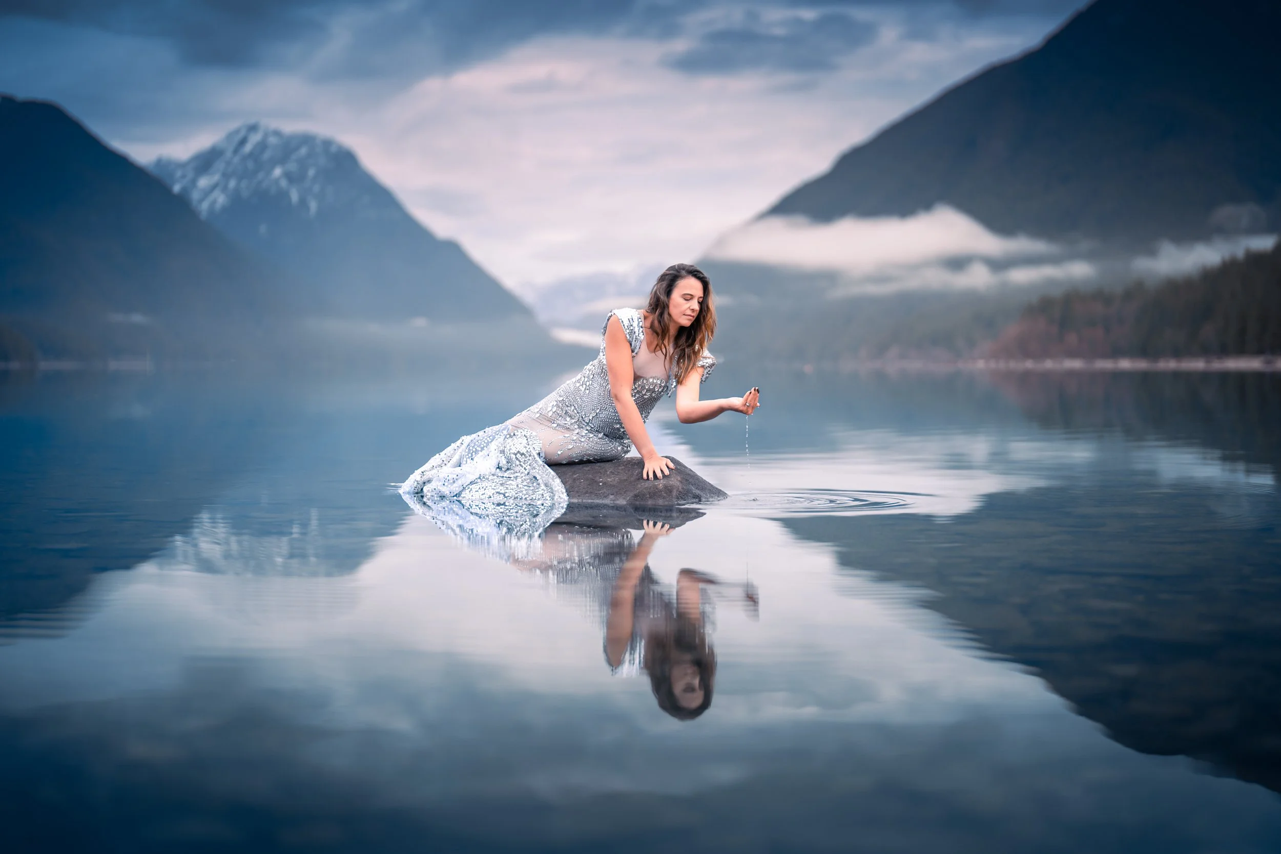 A woman in a sparkly gray dress kneels on a rock in a calm lake, with mountains and clouds in the background, as she dips her hand into the water.