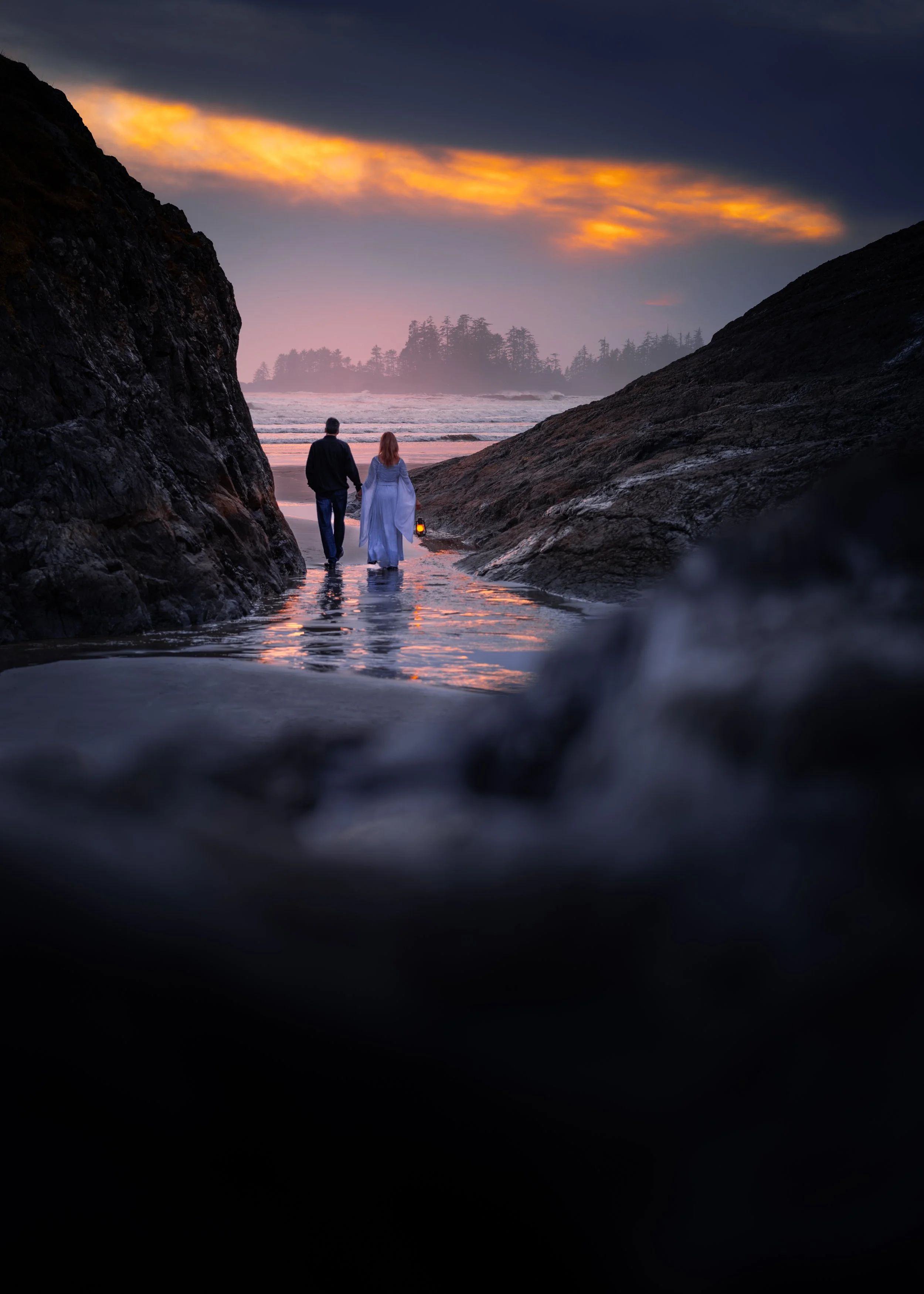 A man and woman walking along a beach at sunset, surrounded by rocks, with the ocean and a distant tree-covered island in the background.
