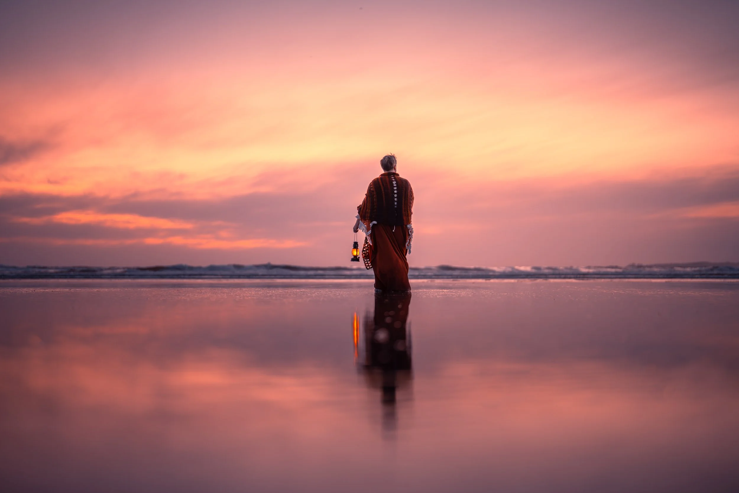 Person in a long dress holding a lantern, walking on a reflective beach during sunset with pink and orange sky.