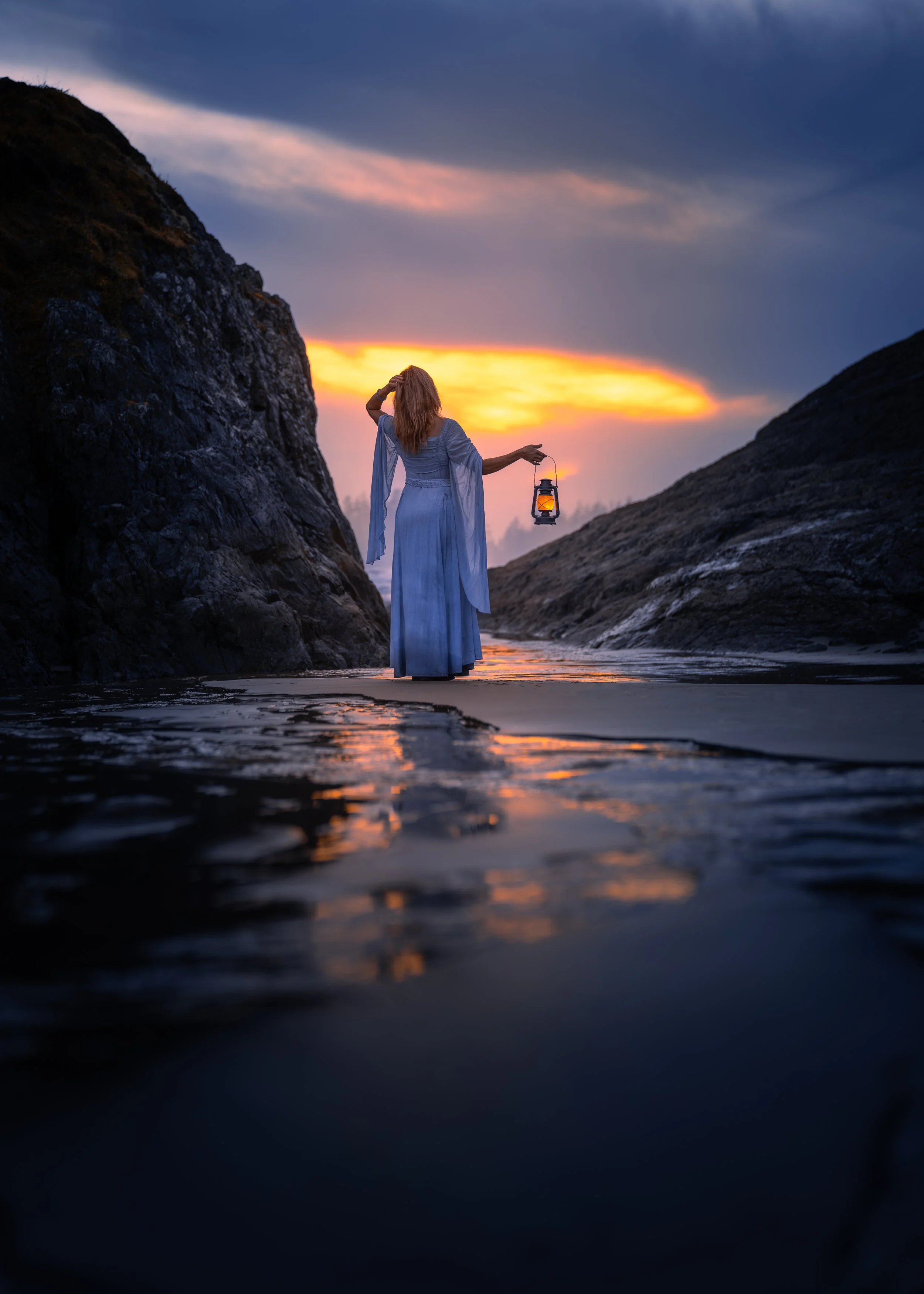 A woman in a flowing blue dress walking in water between rocky cliffs at sunset, holding a lantern.
