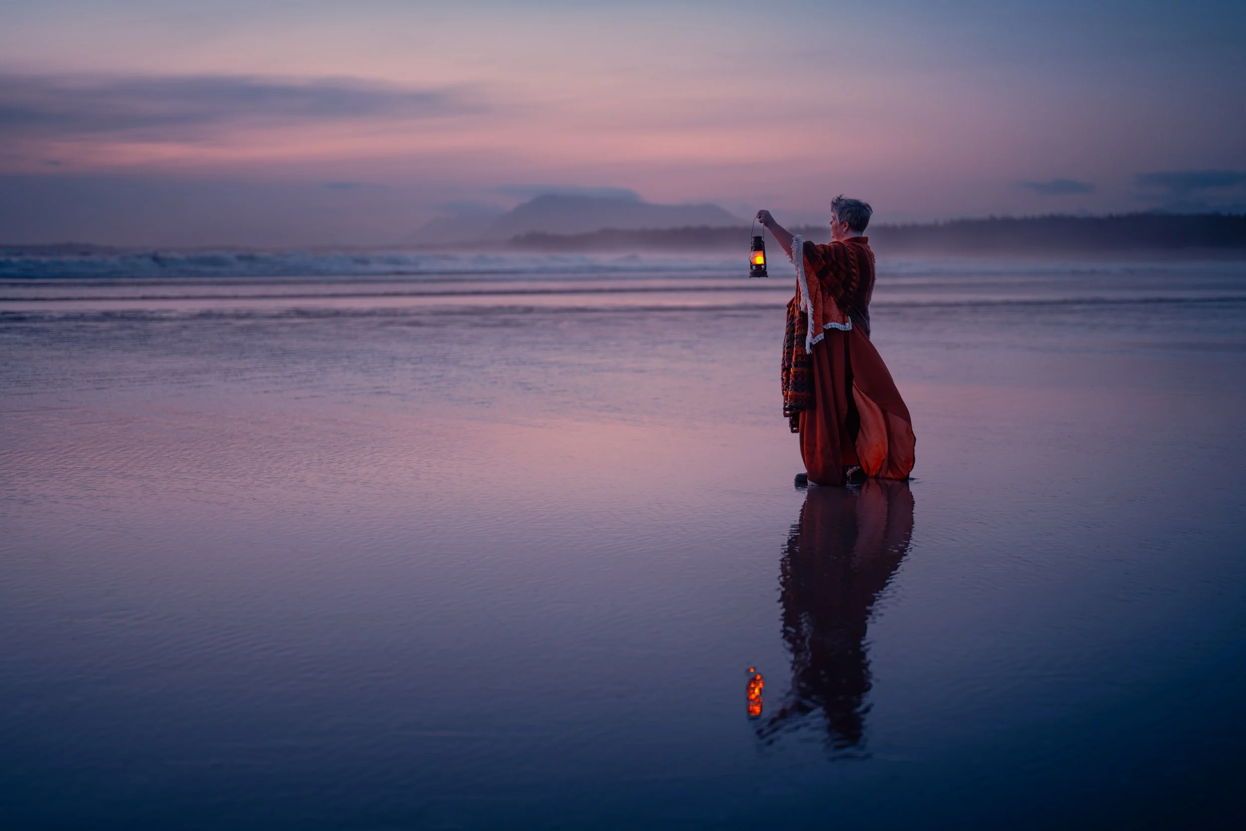 A person standing in shallow water at the beach during sunset, holding a lantern, with the sky reflecting pink and purple hues and distant mountains in the background.