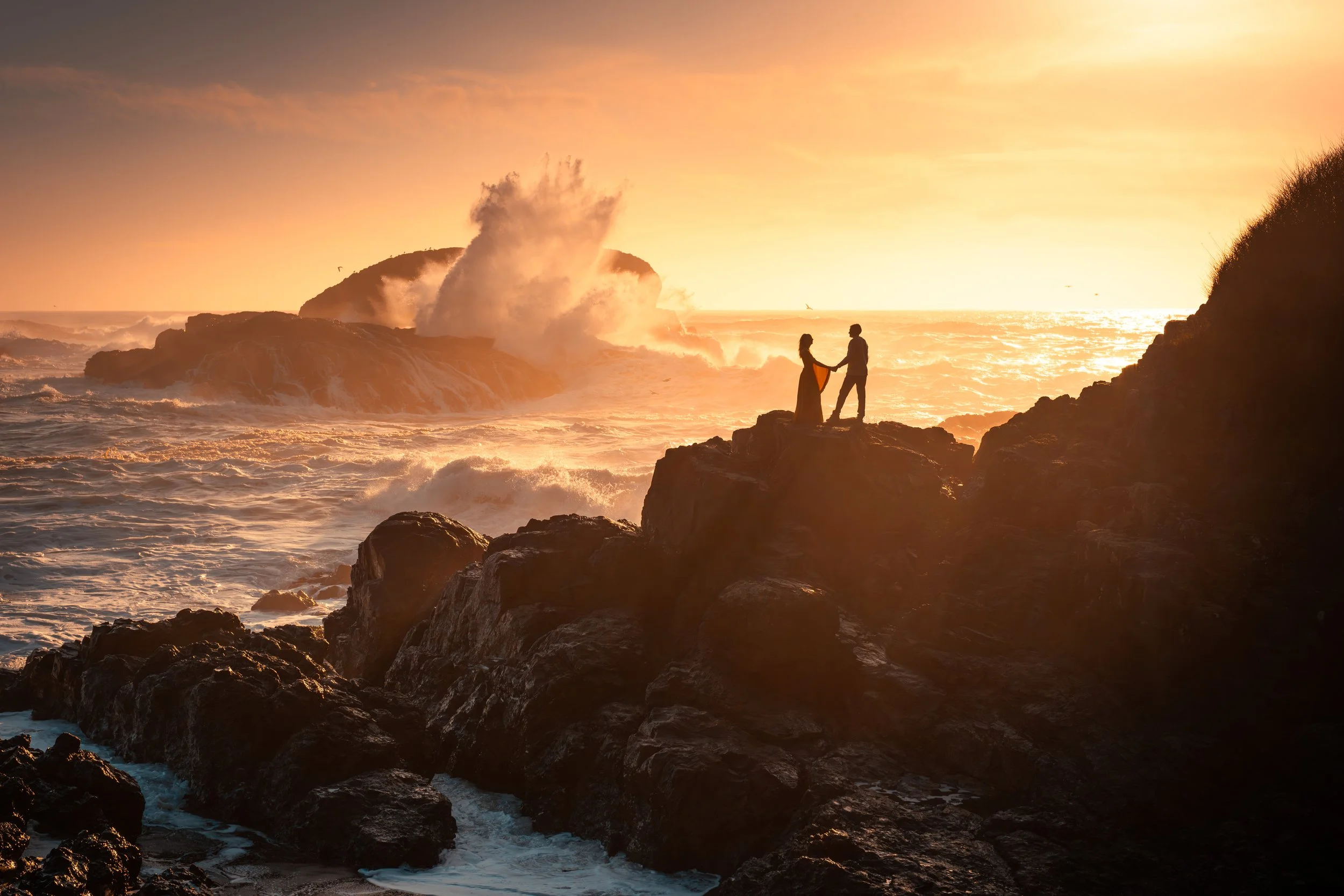 A couple holding hands on rocks by the ocean during sunset with waves crashing behind them.