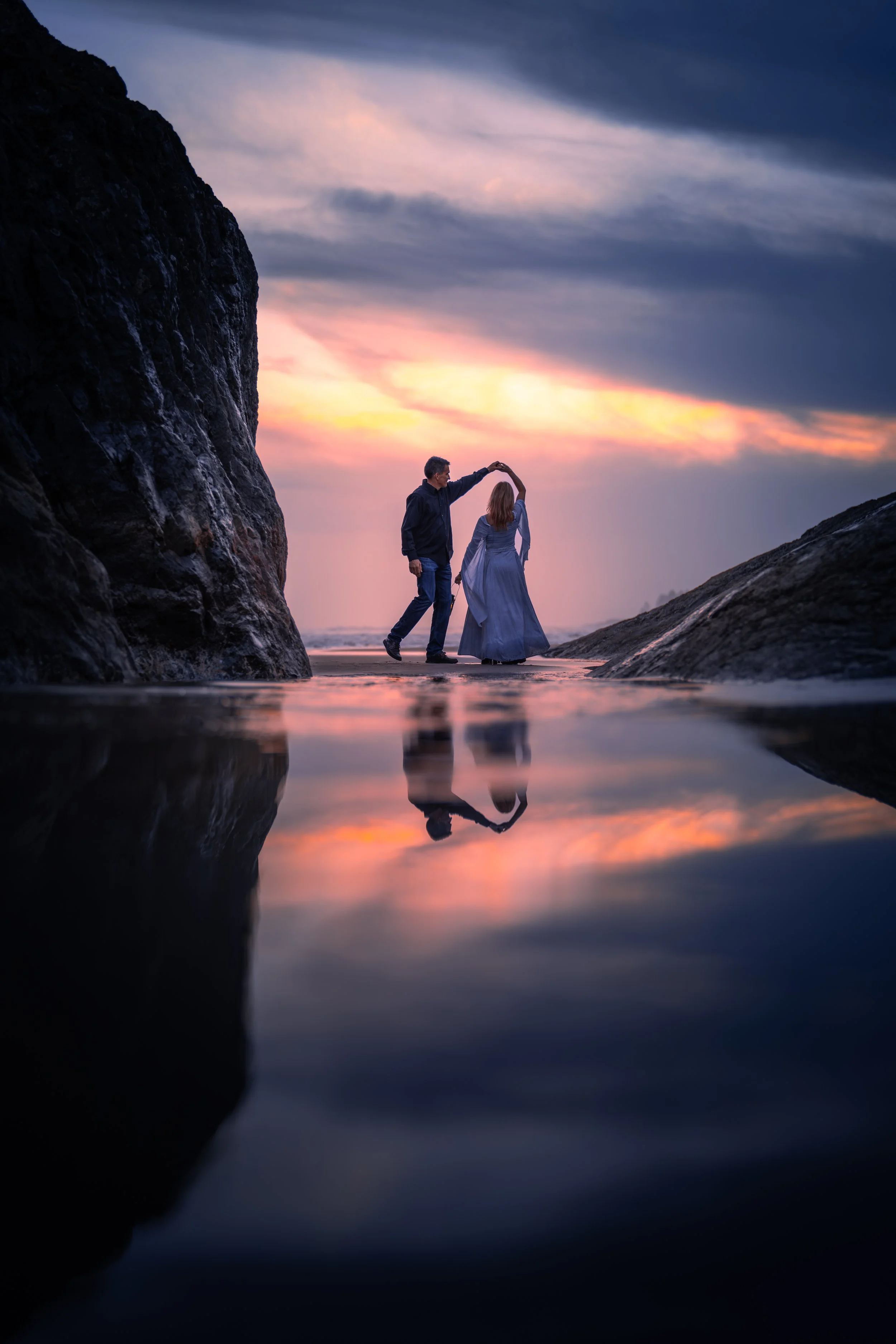 A man and woman dancing on the beach at sunset, with rocks on either side and the sky reflected in the wet sand.