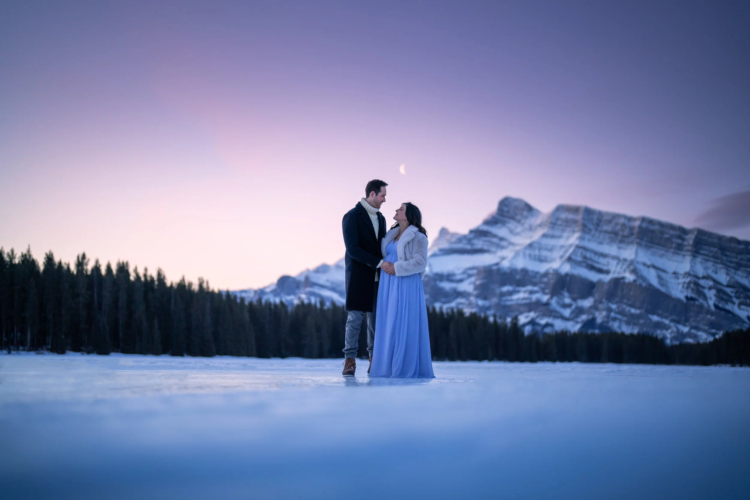 A couple stands closely together on a snowy landscape with a mountain range and a twilight sky in the background.