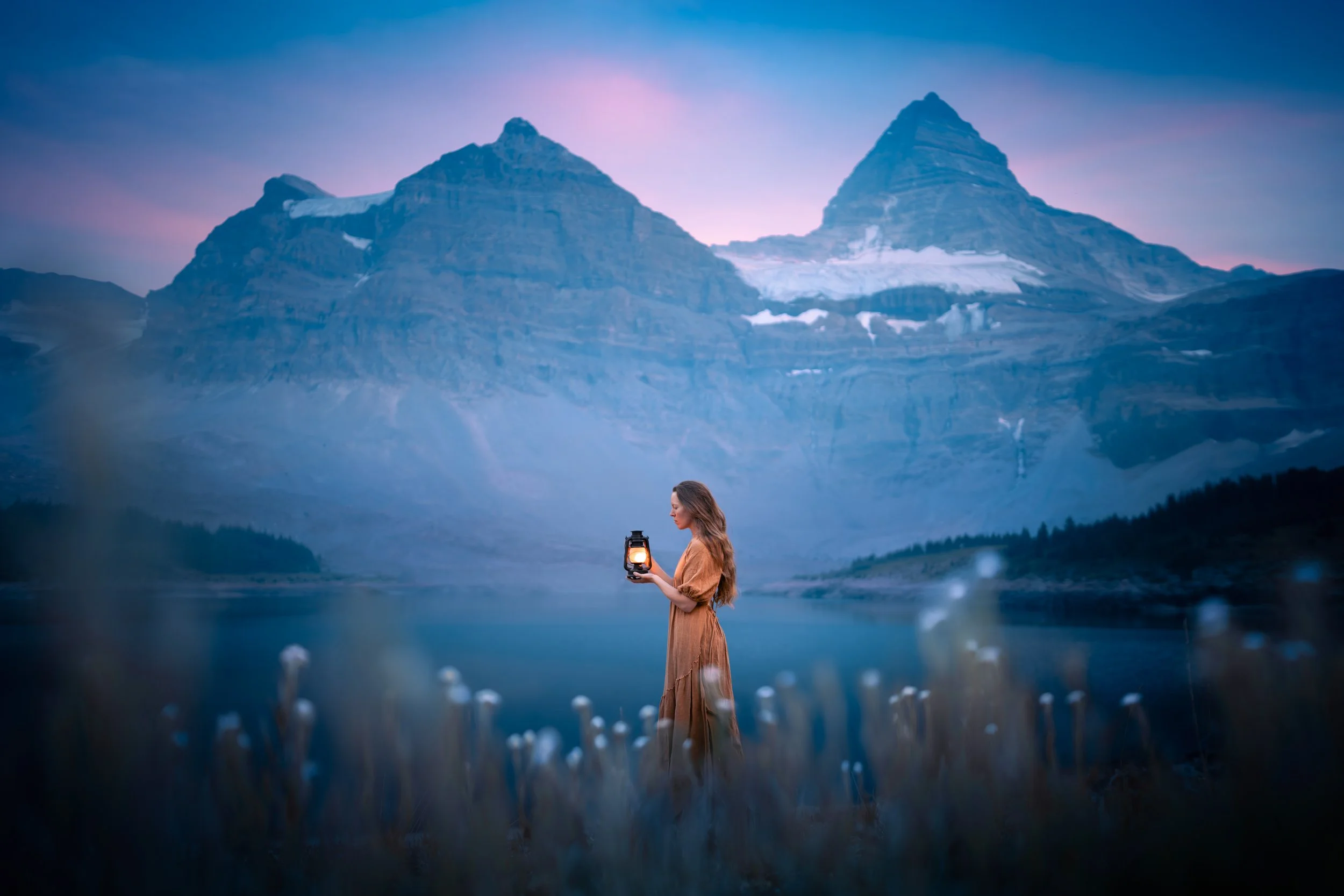 A woman in a brown dress holding a lantern during twilight in a mountainous landscape with a lake and tall peaks in the background.