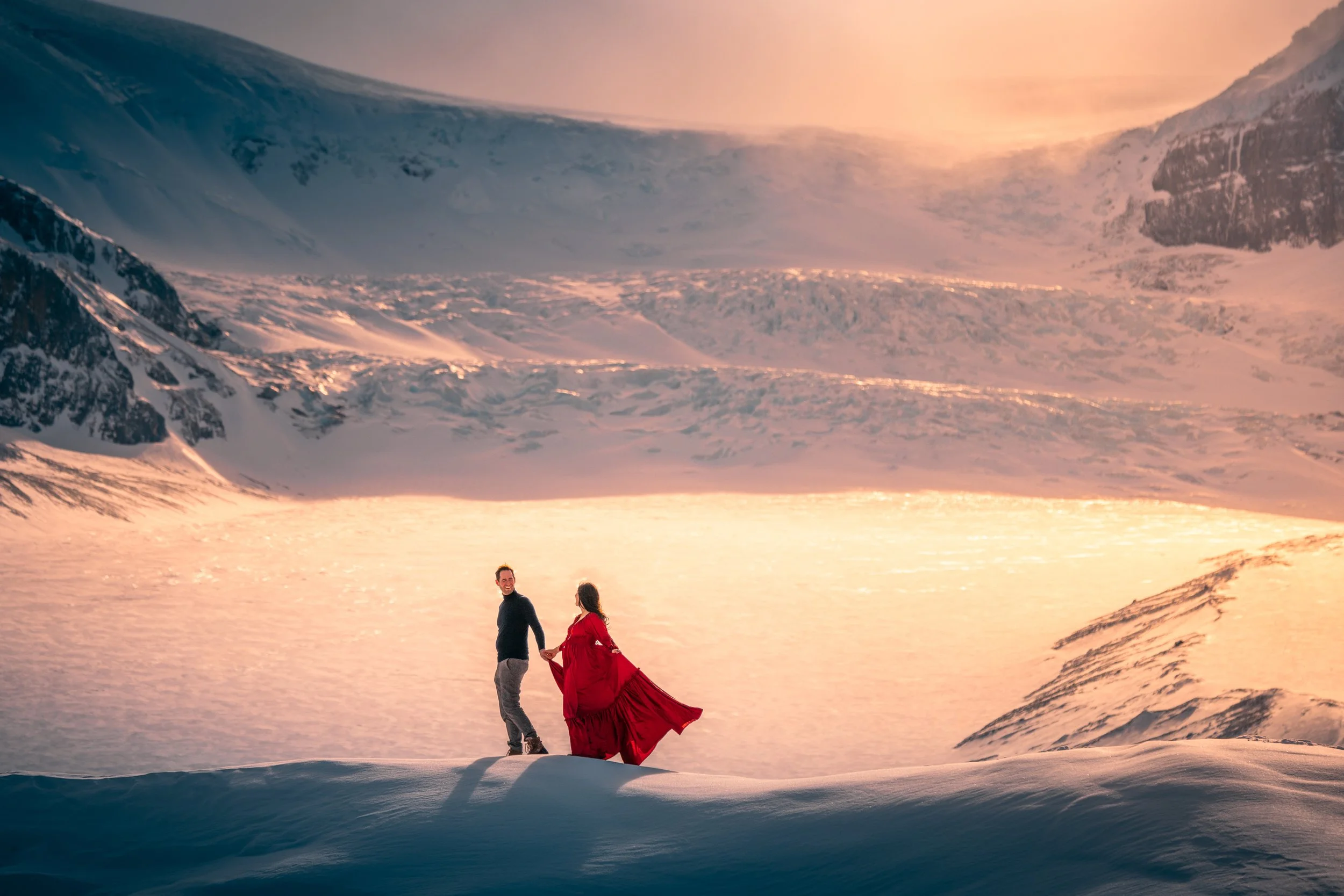 A couple standing in a snowy landscape at sunset with mountains in the background. The woman is wearing a long red dress, and the man is dressed in dark clothing.