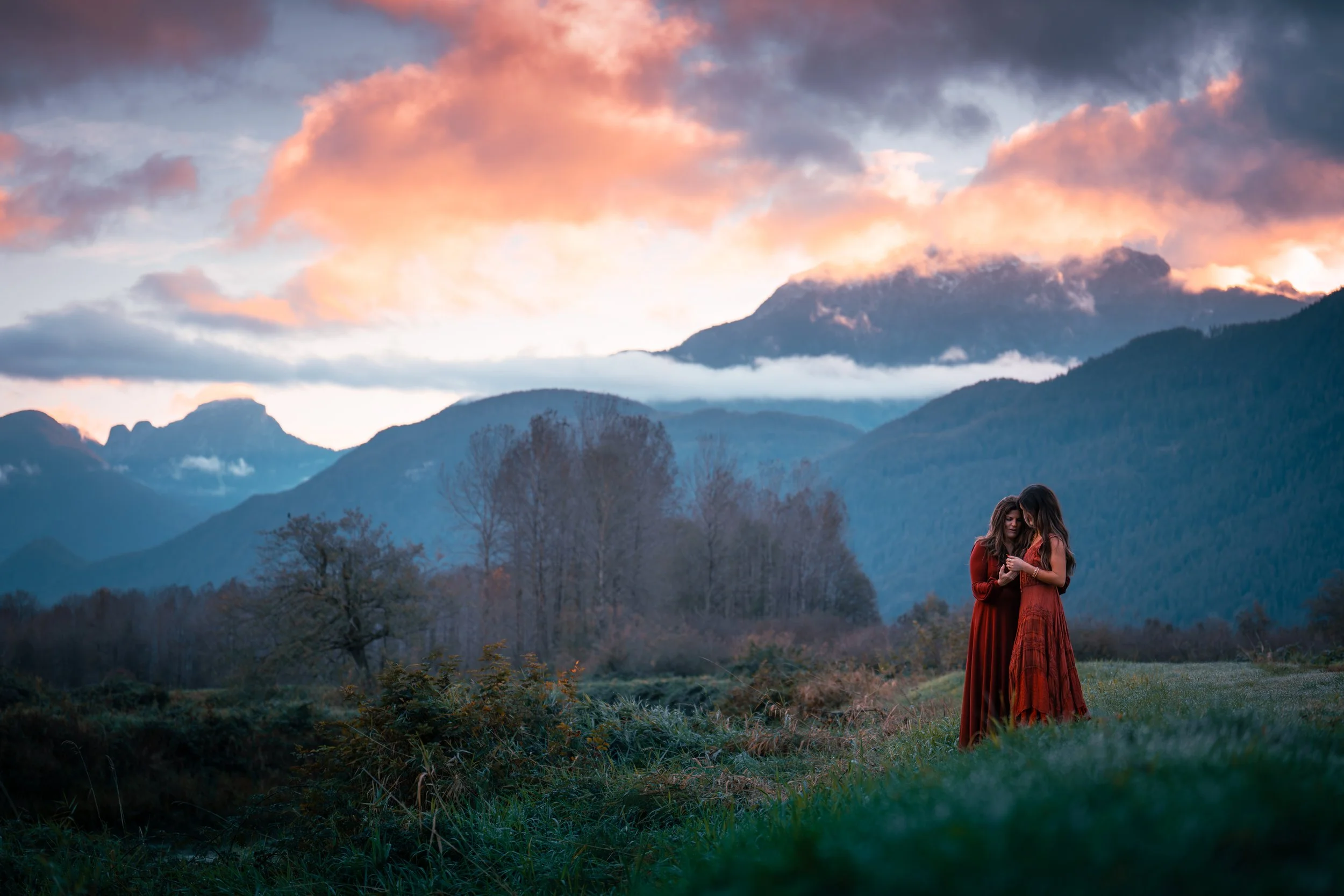Two women in red dresses standing in a grassy field with mountains and a colorful sky with clouds at sunset or sunrise in the background.