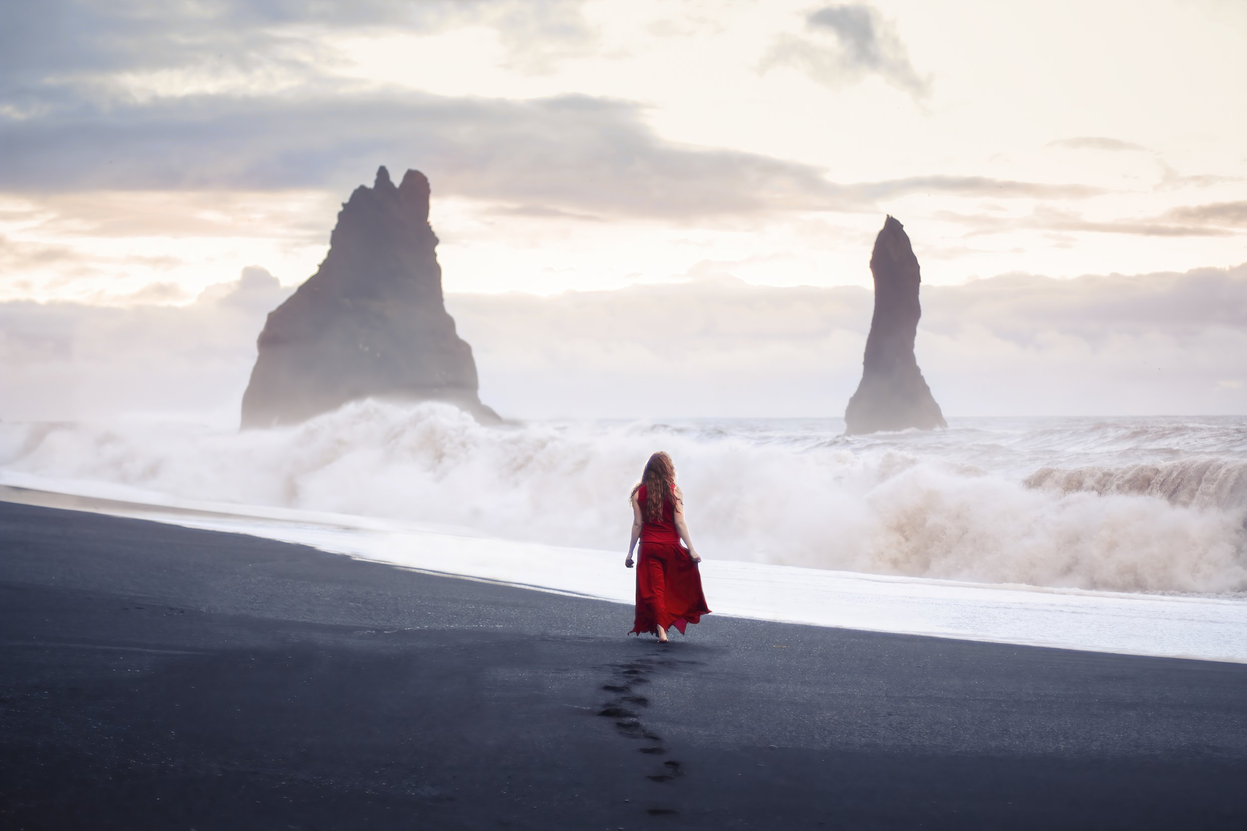 A woman in a red dress walking along a black sand beach towards the ocean with large waves crashing. Sea stacks are visible in the distance under a cloudy sky.