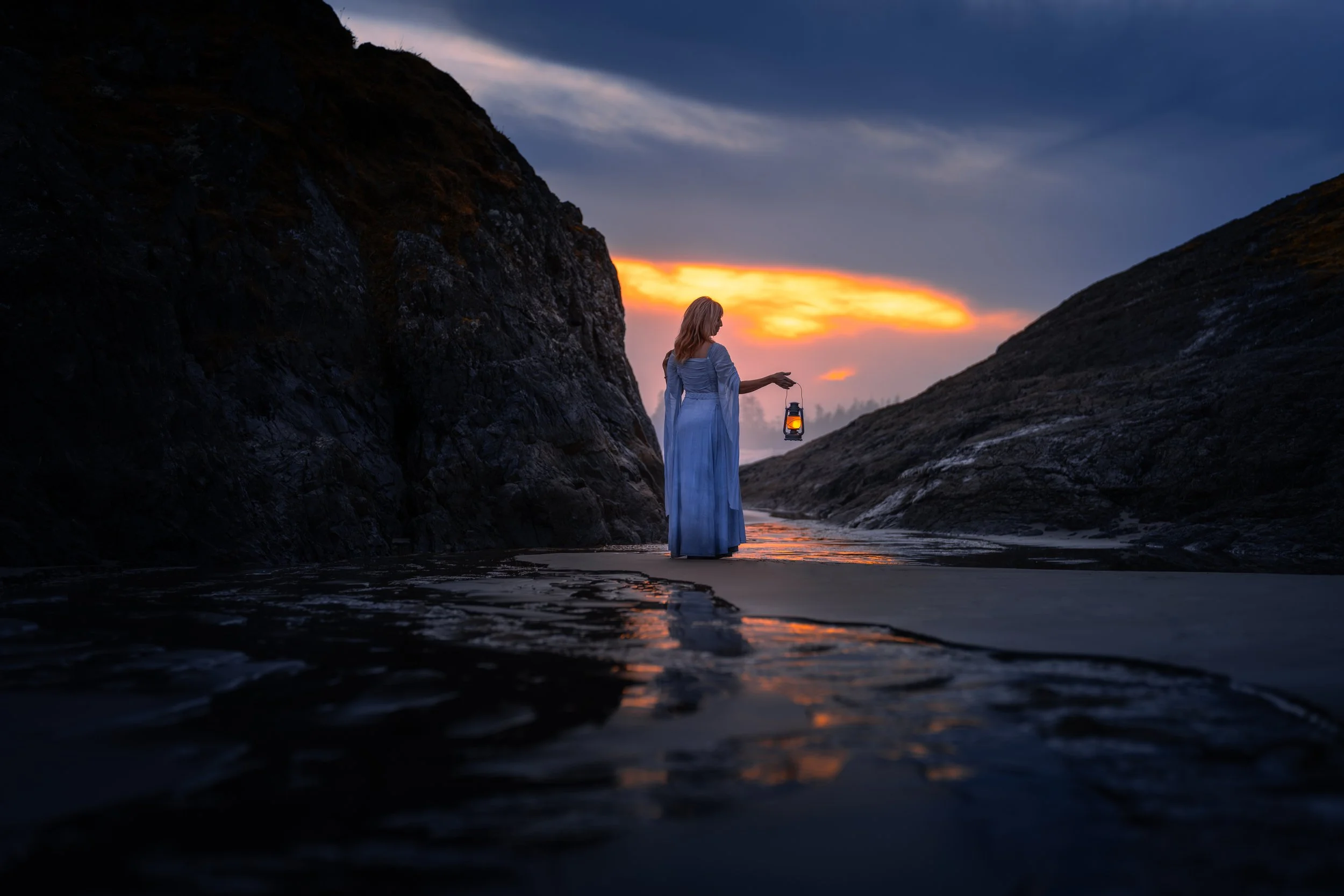A woman in a long blue dress holding a lantern, standing in shallow water between two rocky cliffs at sunset.