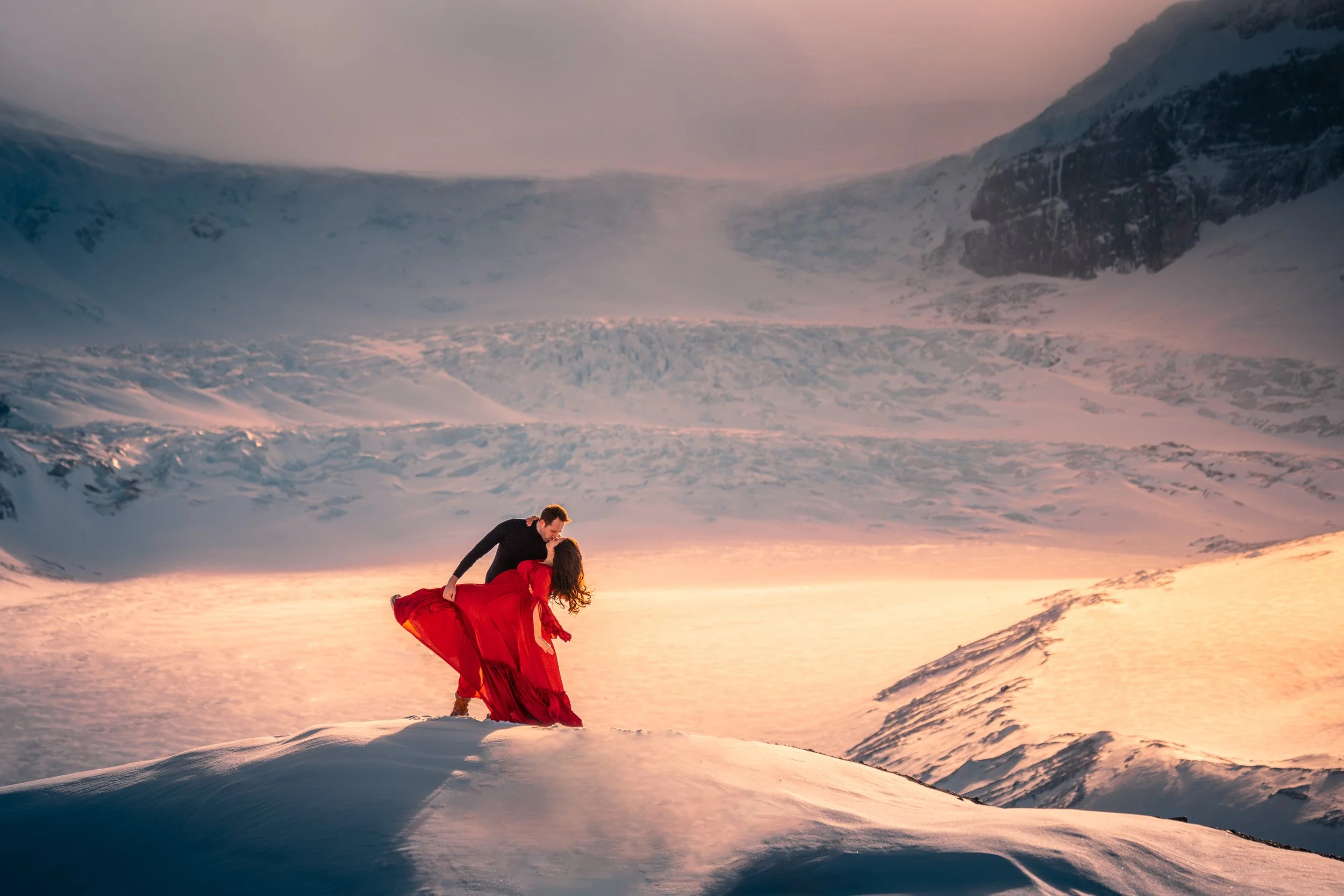 A couple dancing on snow-covered terrain with mountains and a colorful, cloudy sky in the background.