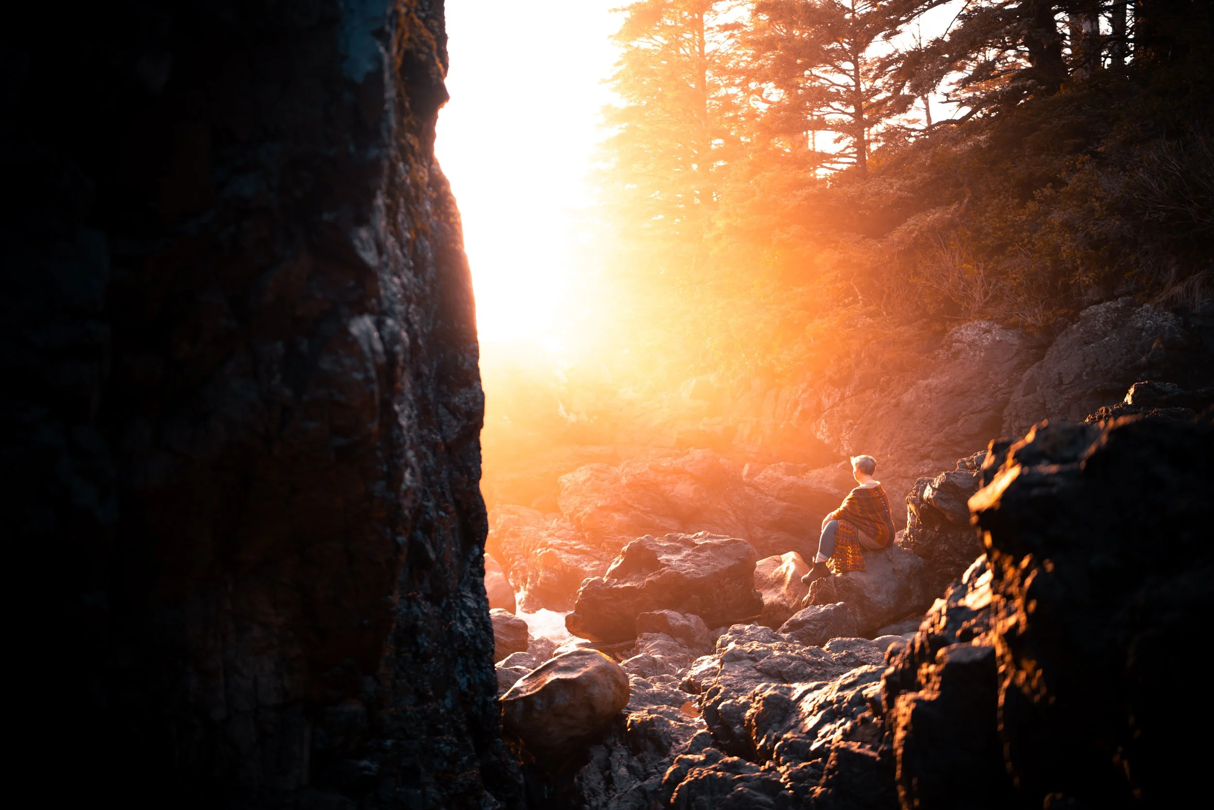 A person sitting on rocks by a river at sunset, surrounded by large dark rocks and trees in the background, with sunlight illuminating the scene.