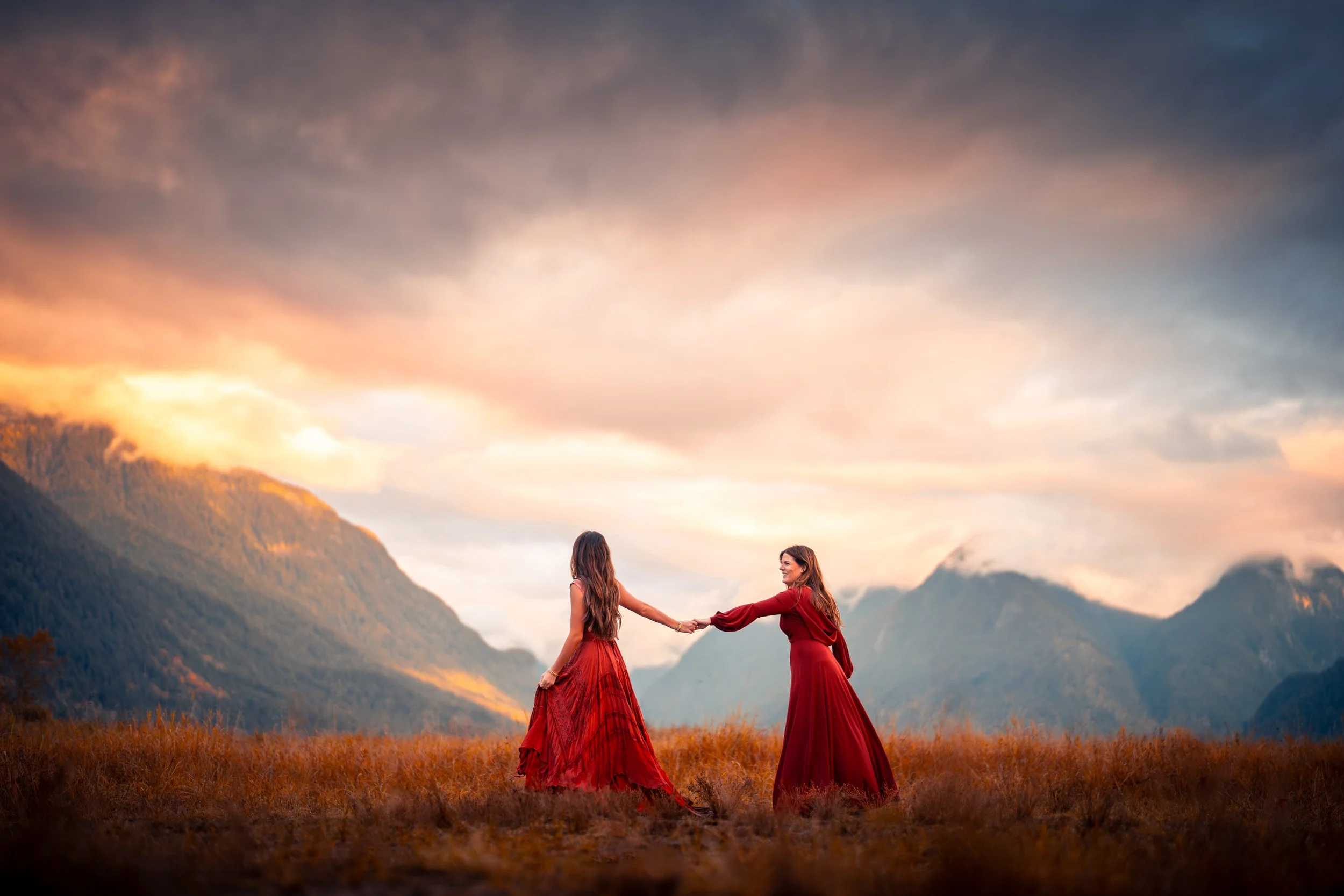 Two women in red dresses holding hands in a field with mountains and a colorful sunset sky in the background.