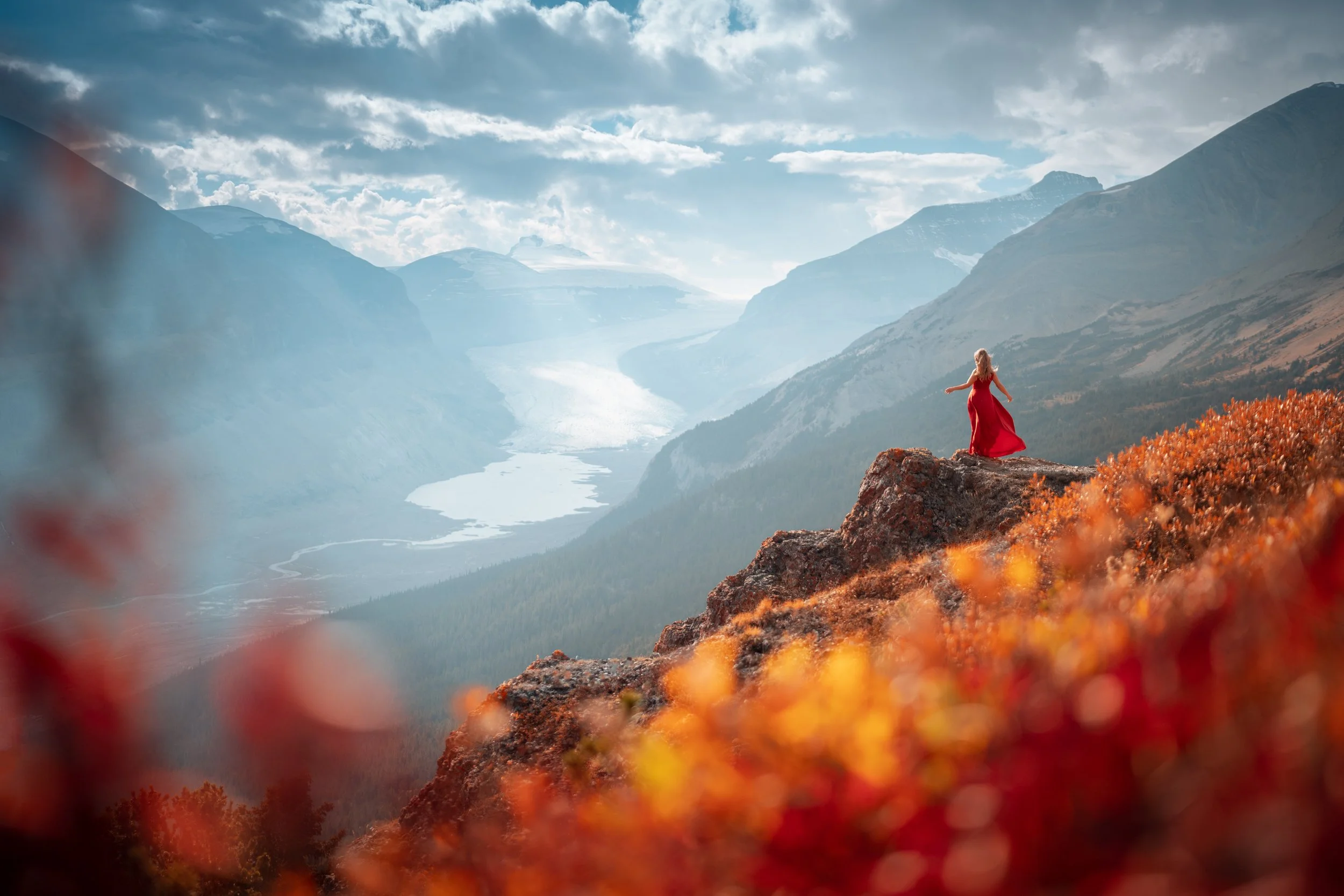 A woman in a red dress standing on a rocky ledge overlooking a mountainous valley with a river, under a partly cloudy sky.