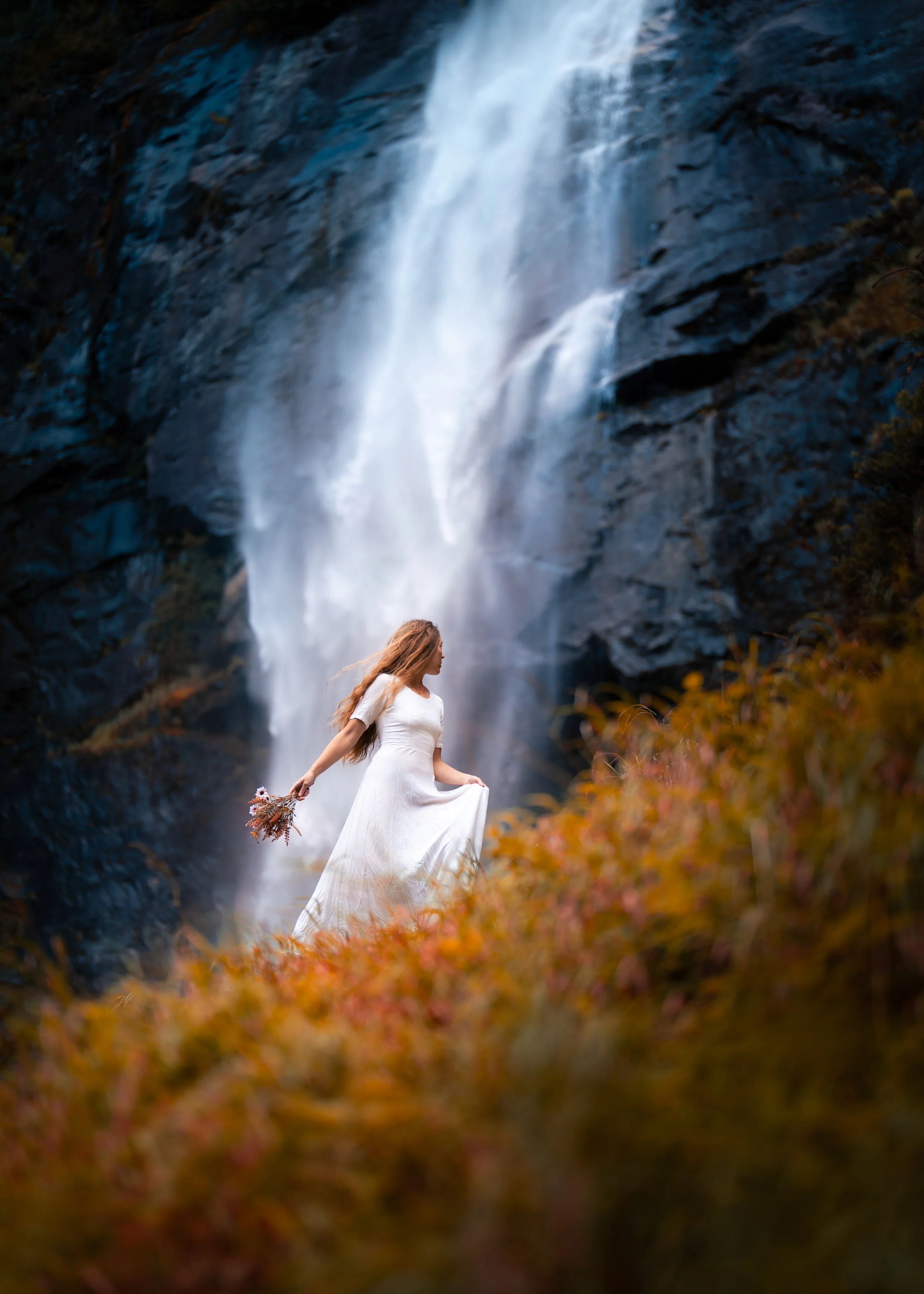A woman in a white dress holding a bouquet stands near a waterfall surrounded by autumn foliage.