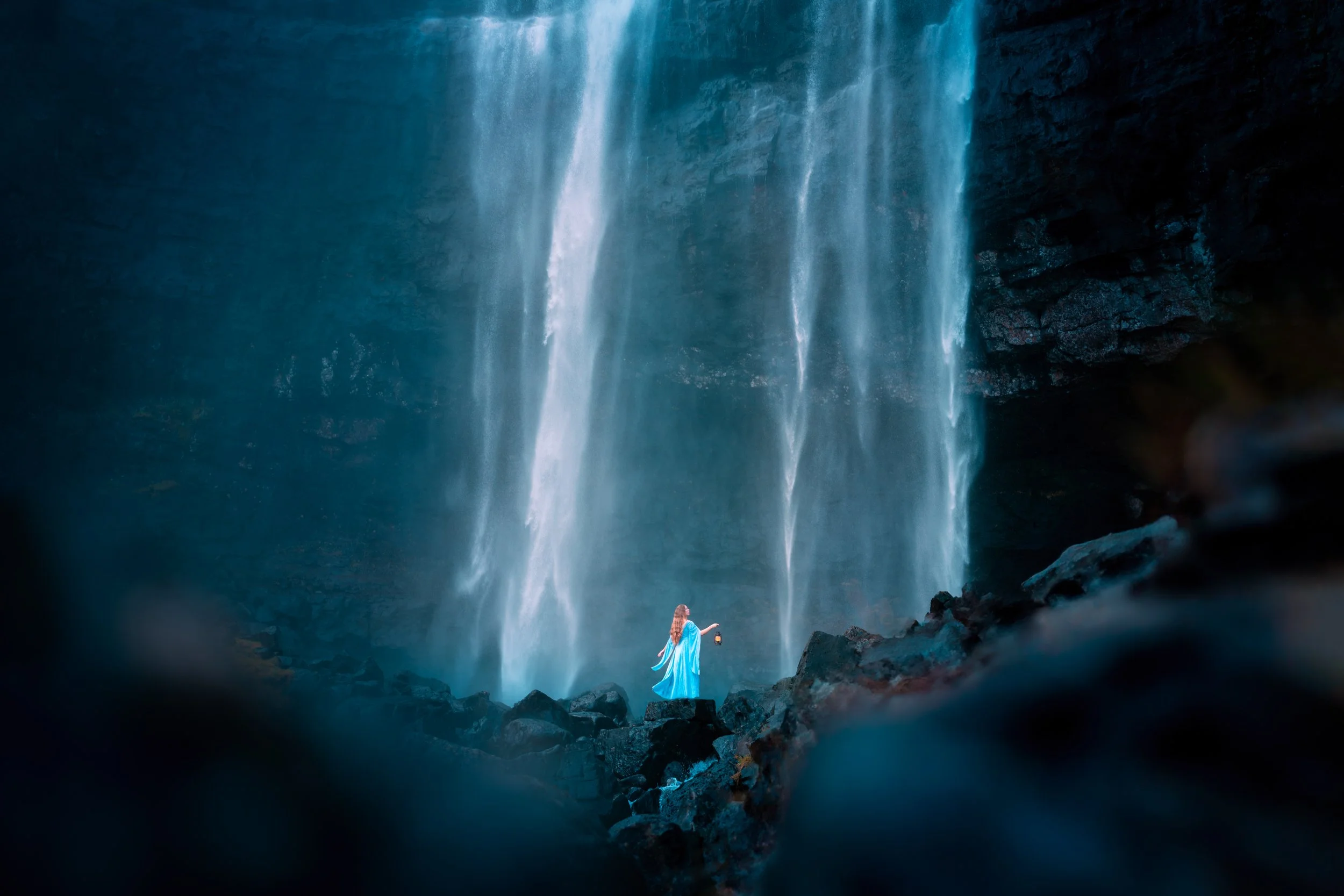 A woman in a flowing blue dress standing on rocks near a tall, cascading waterfall, holding a lantern.