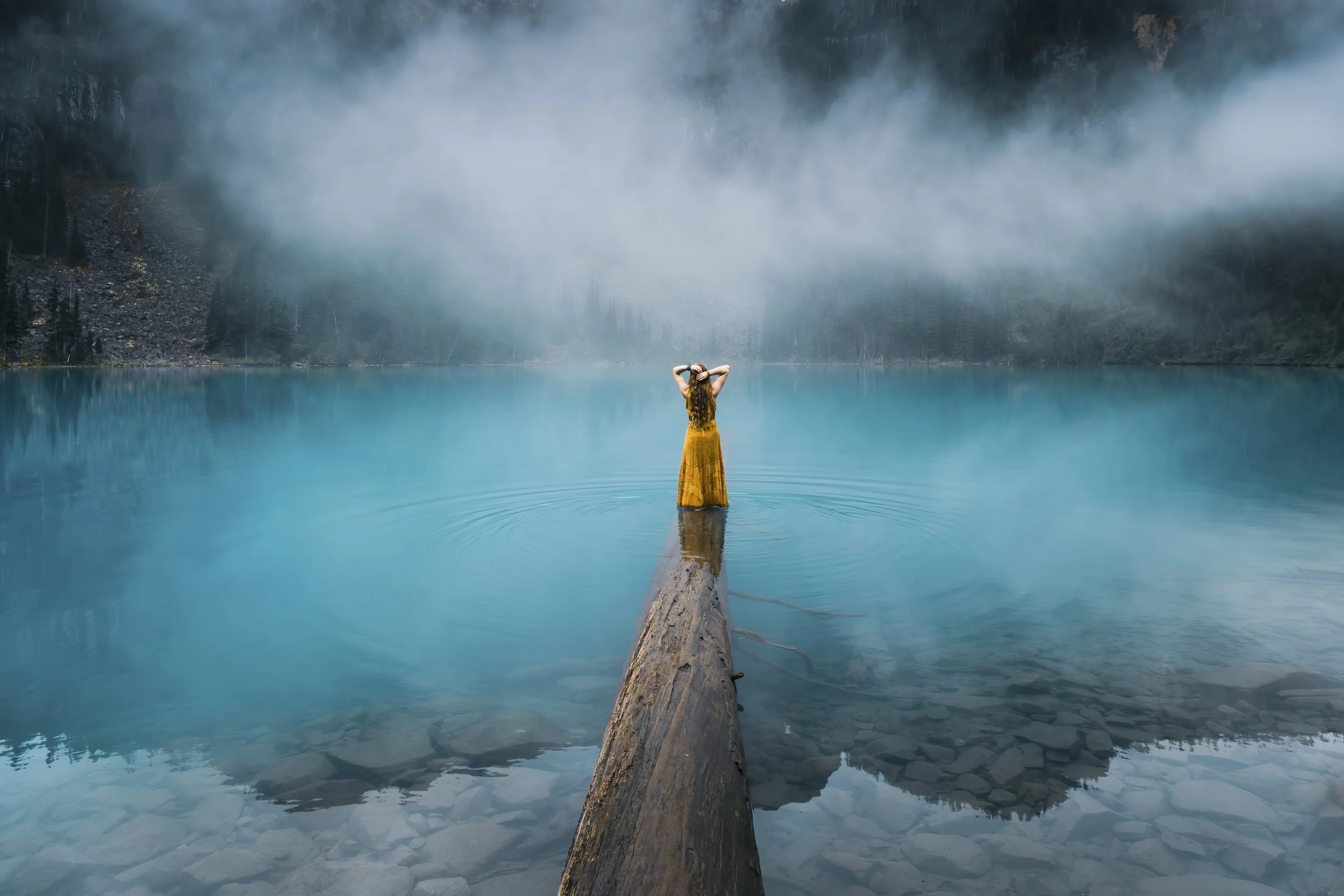 A woman in a yellow dress standing on a log in a calm, blue lake with mountains and fog in the background.