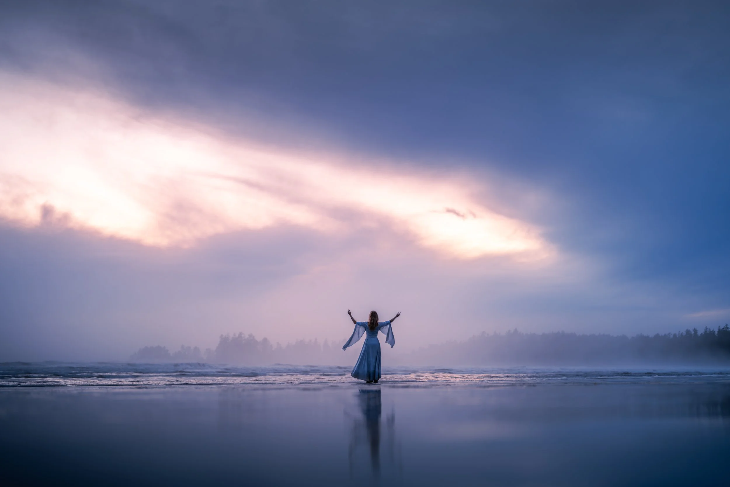 A woman in a flowing dress standing on the wet sand of a beach with arms raised, facing the ocean under a cloudy sky with hint of sunset colors.