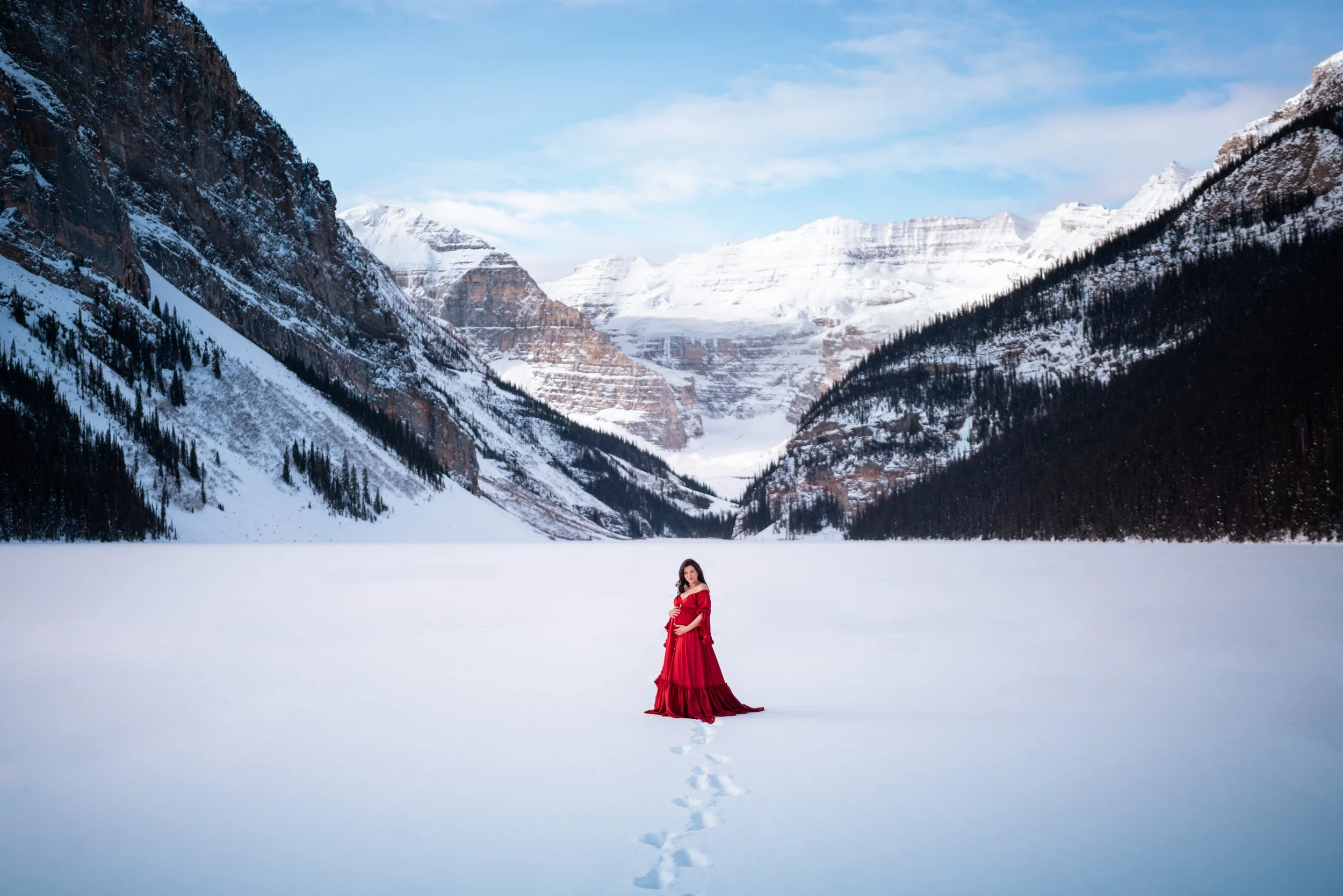 A woman in a red dress standing on snowy landscape with mountains and a frozen lake in the background.