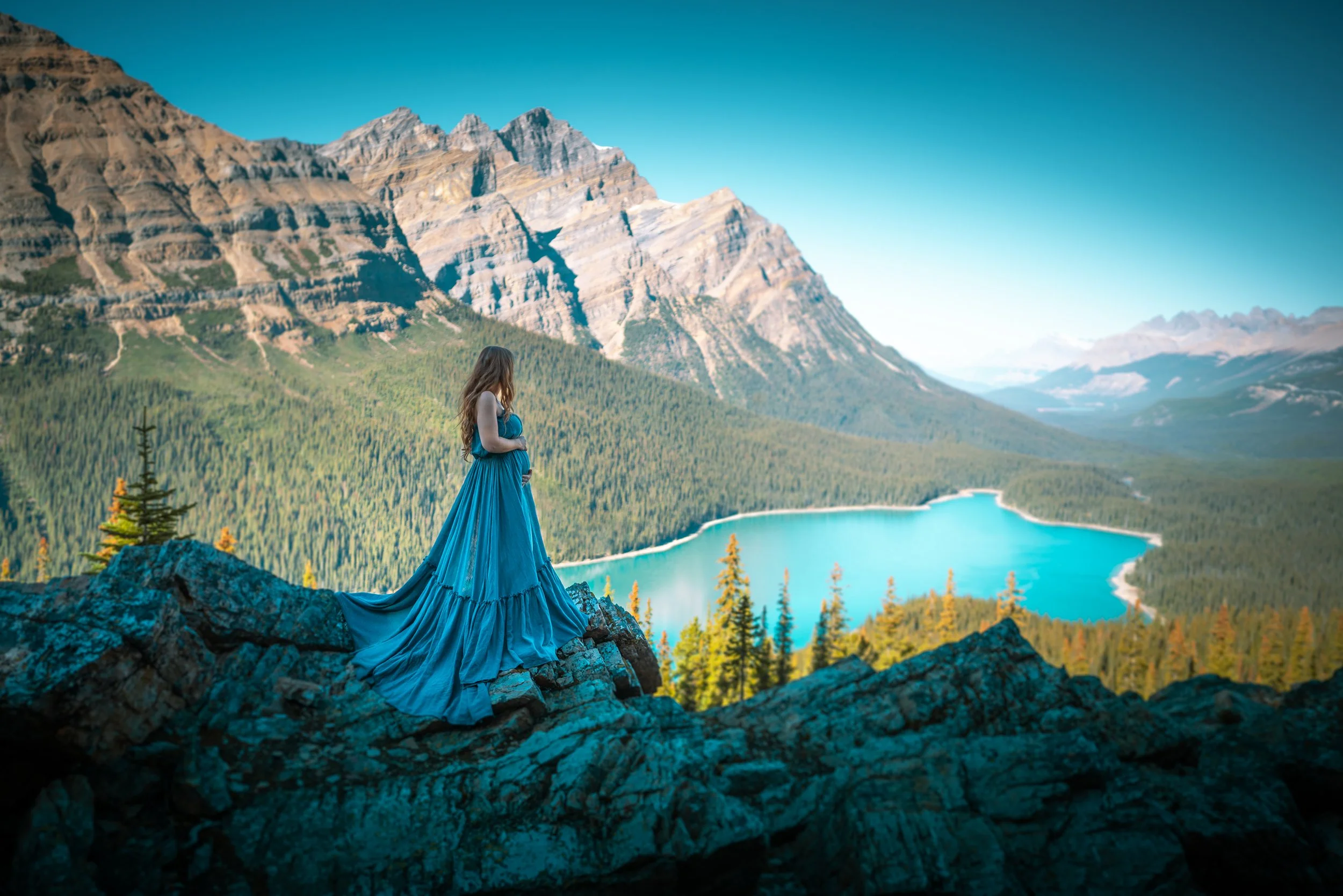 A woman in a long blue dress standing on rocks overlooking a lake with mountains in the background.