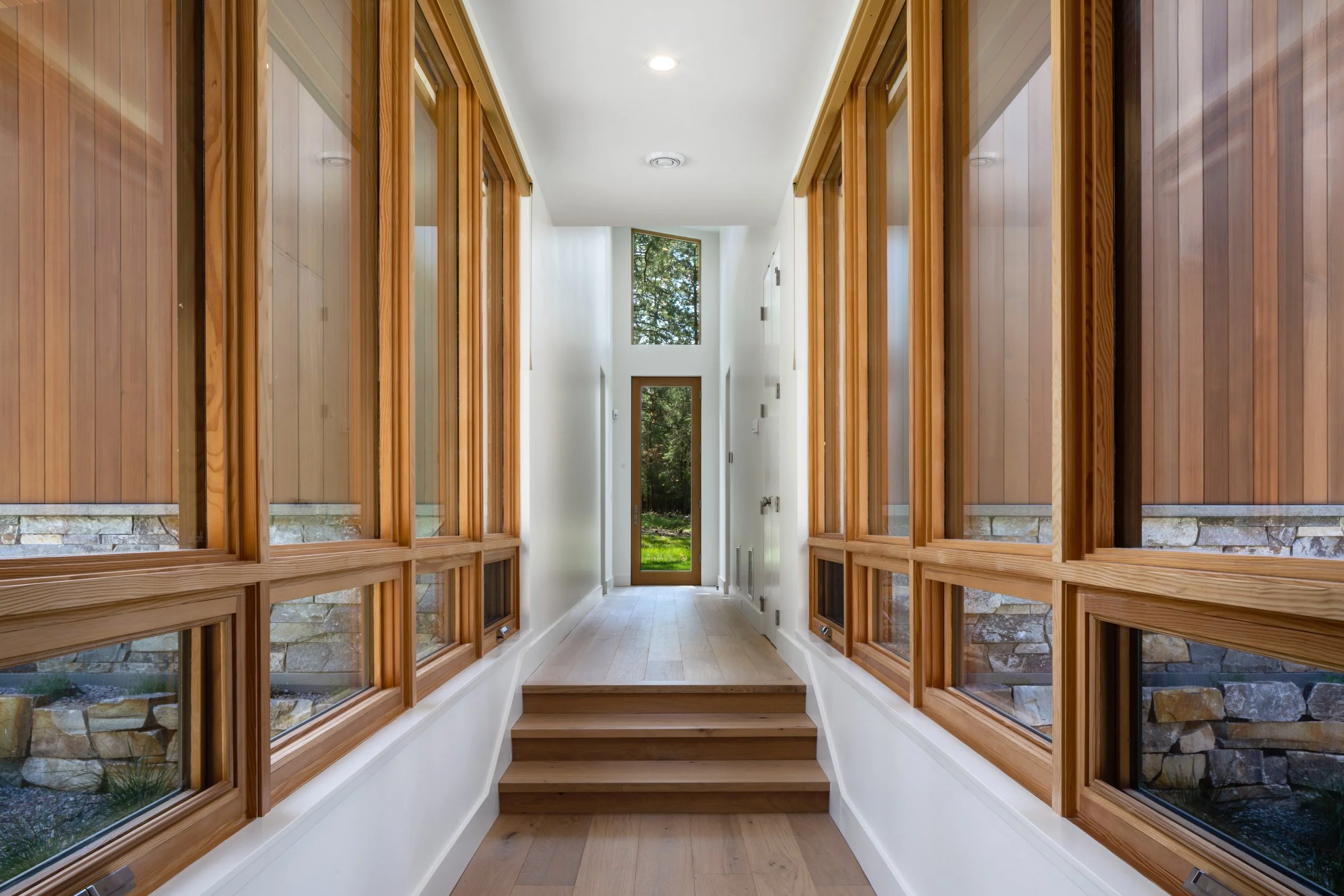 Bright hallway with large windows on both sides featuring wood framing, leading to a door with glass panels and a view of greenery outside.