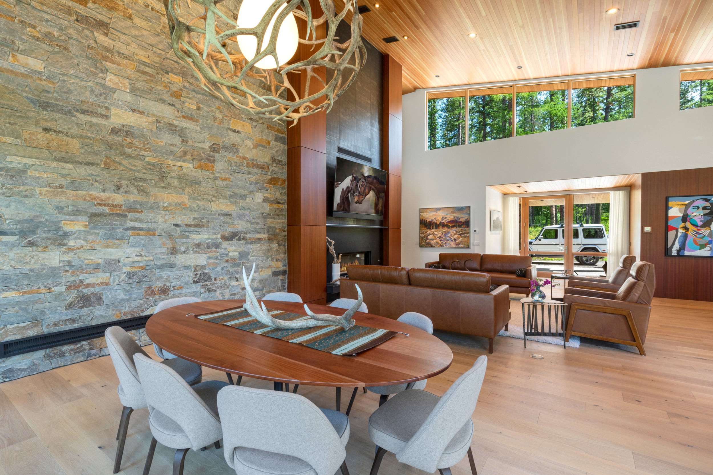 Living room with stone accent wall that was completed by Lindberg Masonry, wooden ceiling, large windows, and modern furniture including a brown leather sofa and armchairs, a round dining table with chairs, and colorful artwork on the walls.