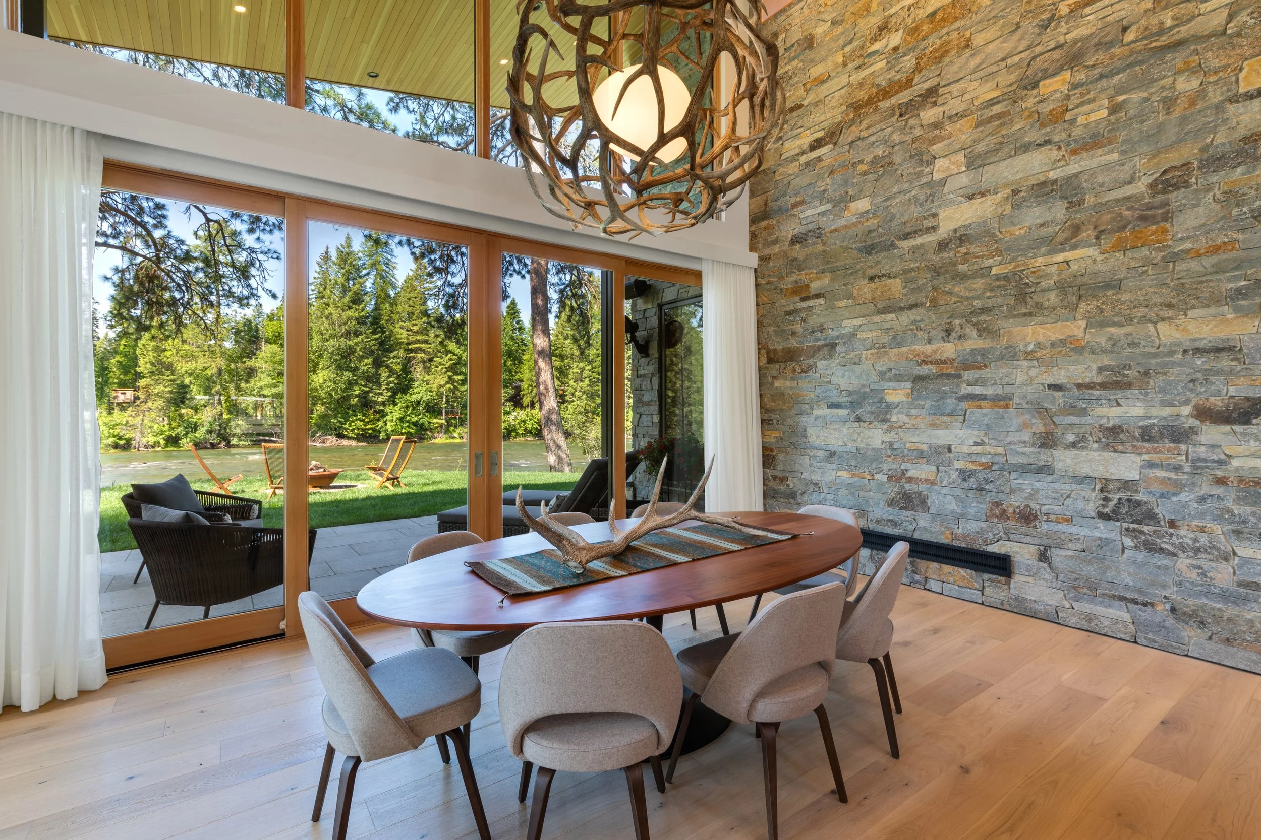Dining room with a wooden oval table, beige chairs, antler centerpiece, large glass sliding doors revealing outdoor patio and greenery, stone accent wall that was done by Lindberg Masonry in Montana, and a rustic chandelier.