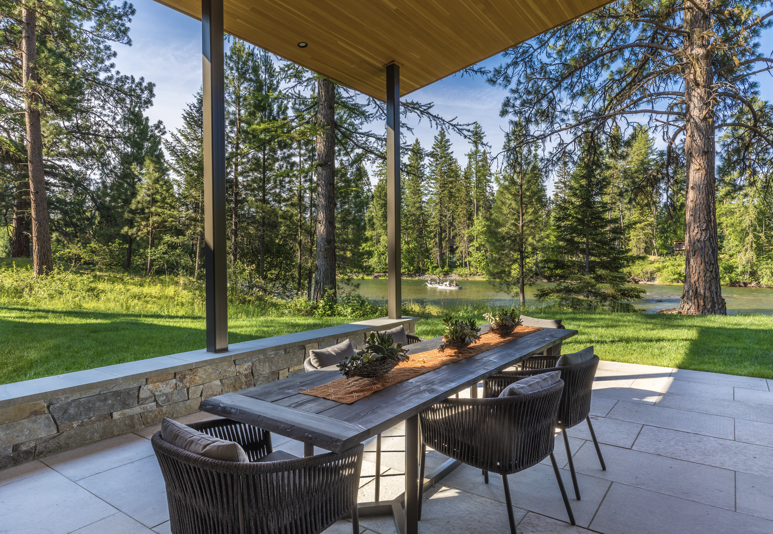 Outdoor patio with rockwork done by Lindberg Masonry a wooden table and six chairs, overlooking a lush green forest and a river with a boat in the distance.