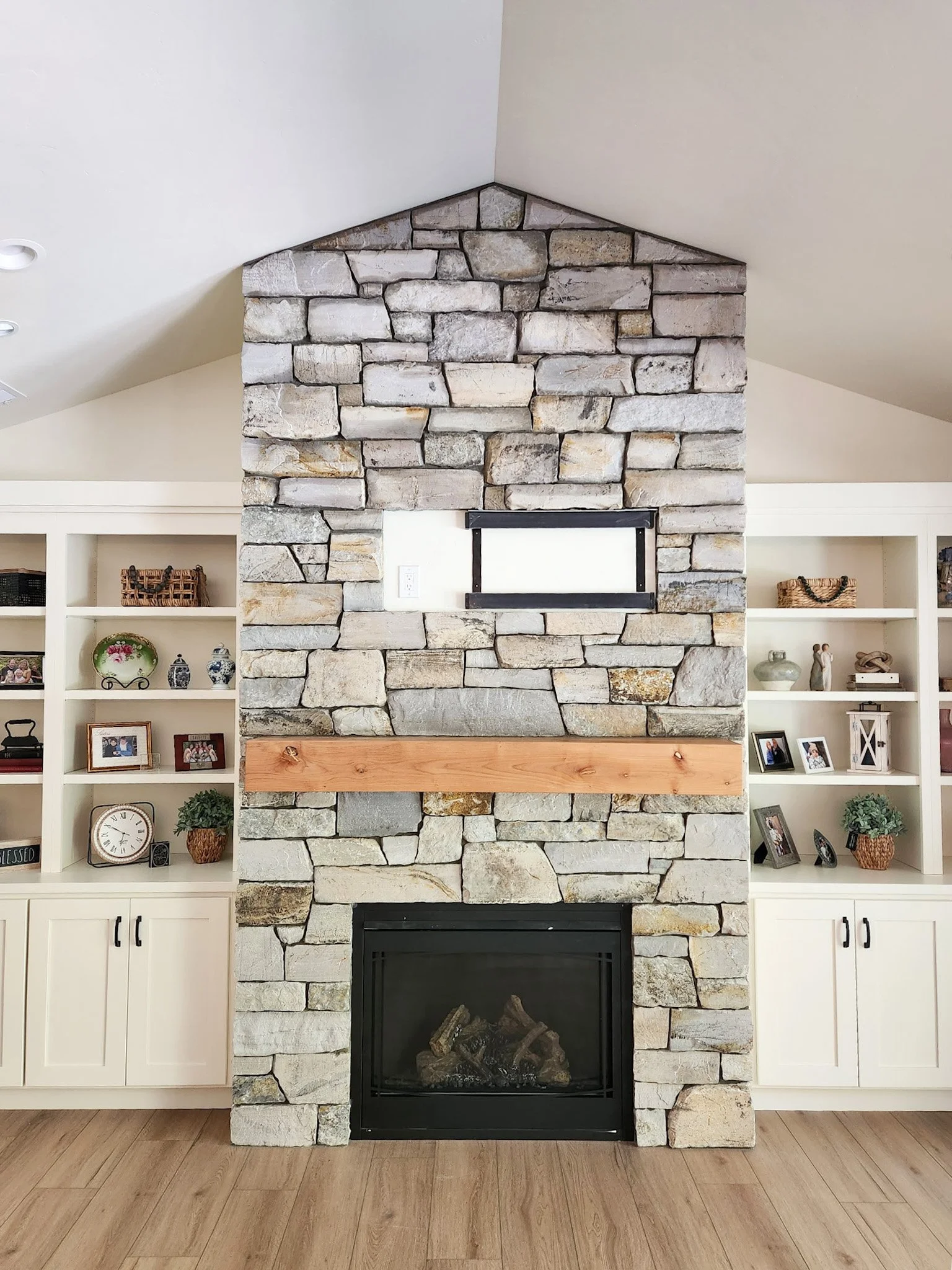 Stone fireplace  done by Lindberg Masonry with wooden mantel in a living room, flanked by built-in white shelves decorated with framed photos, clocks, and ornaments.