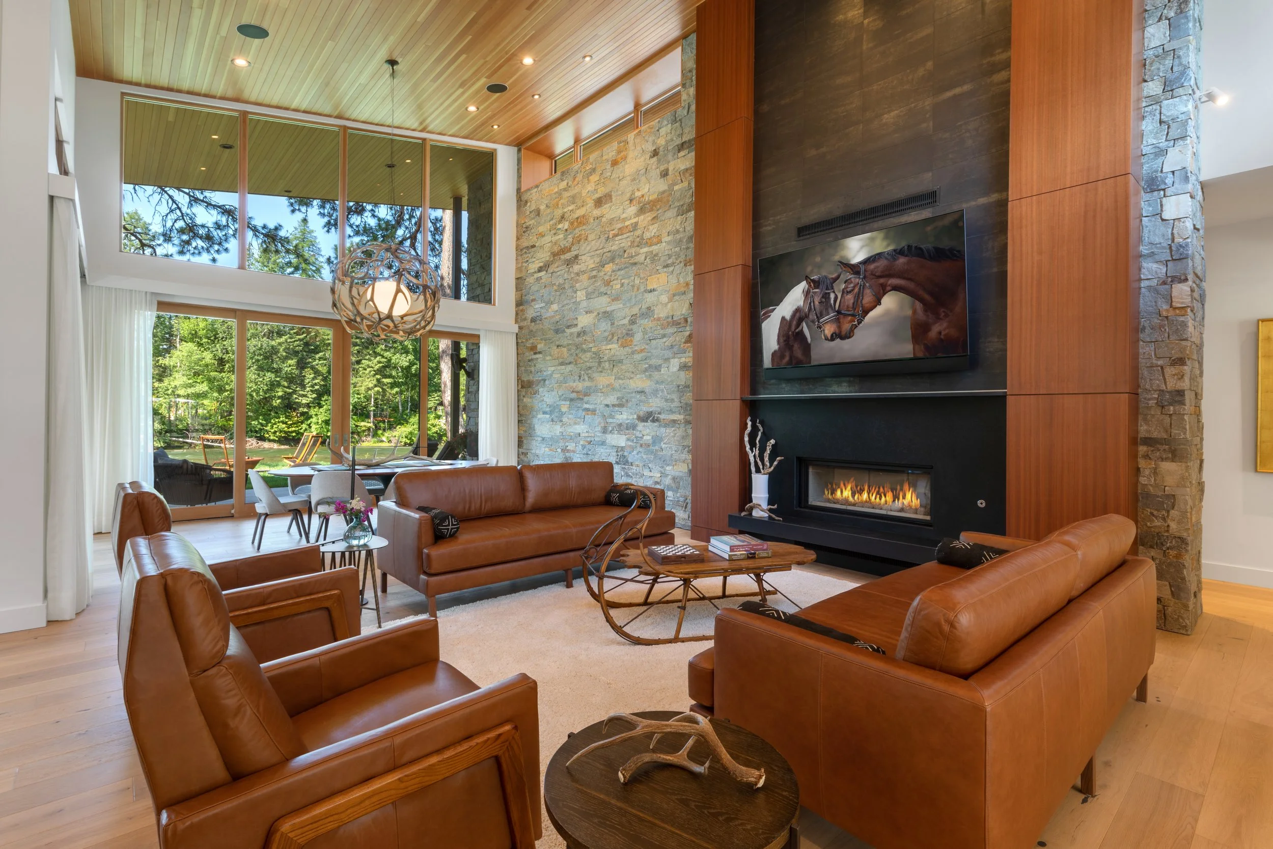Living room with large windows, stone fireplace that was completed by Lindberg Masonry, leather couches, coffee table, TV, and view of outdoor greenery.