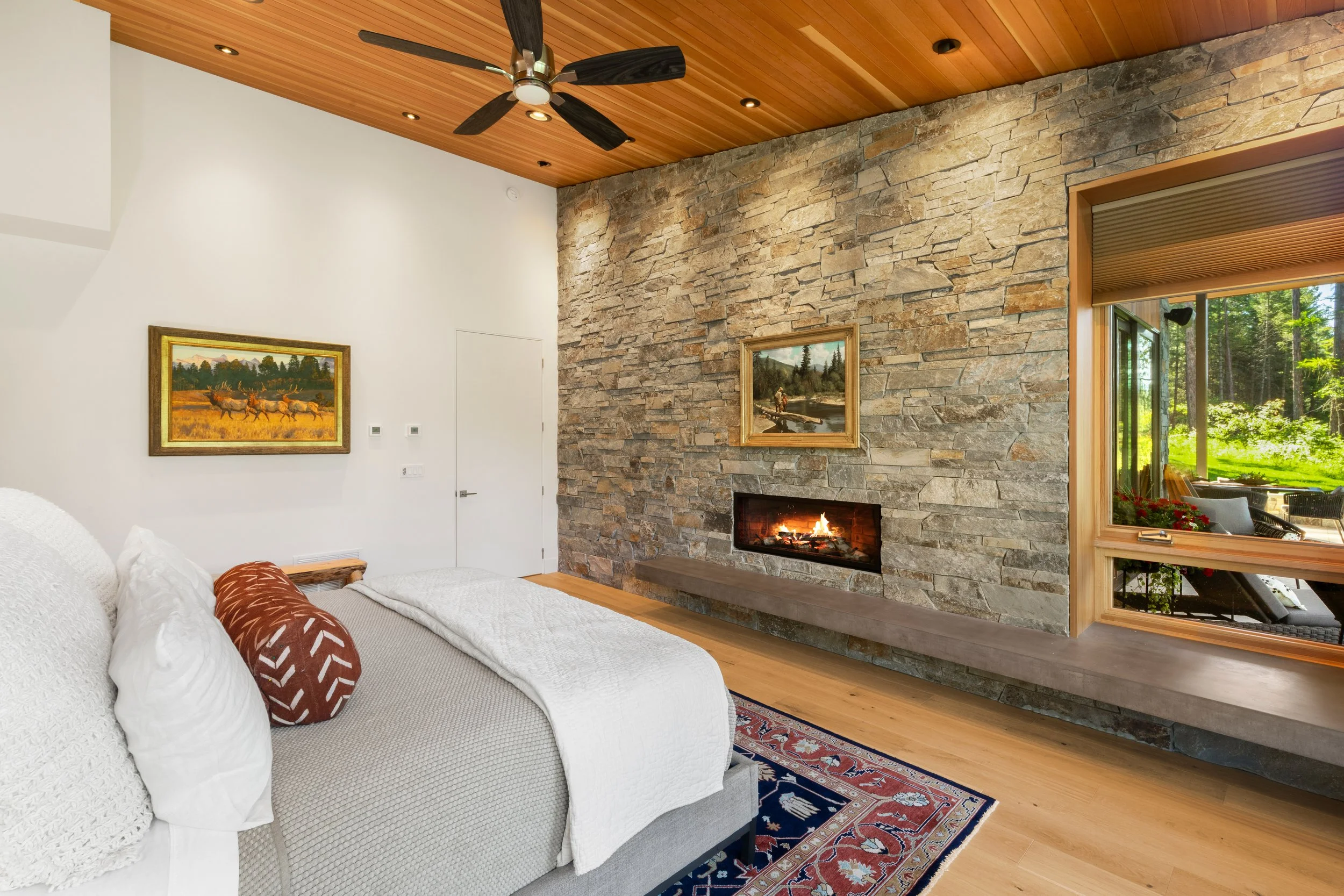 Room with a white bed, stone fireplace, wood ceiling, ceiling fan, paintings, and large window showing outdoor greenery. Stonework completed by Lindberg Masonry in Flathead Valley Montana.