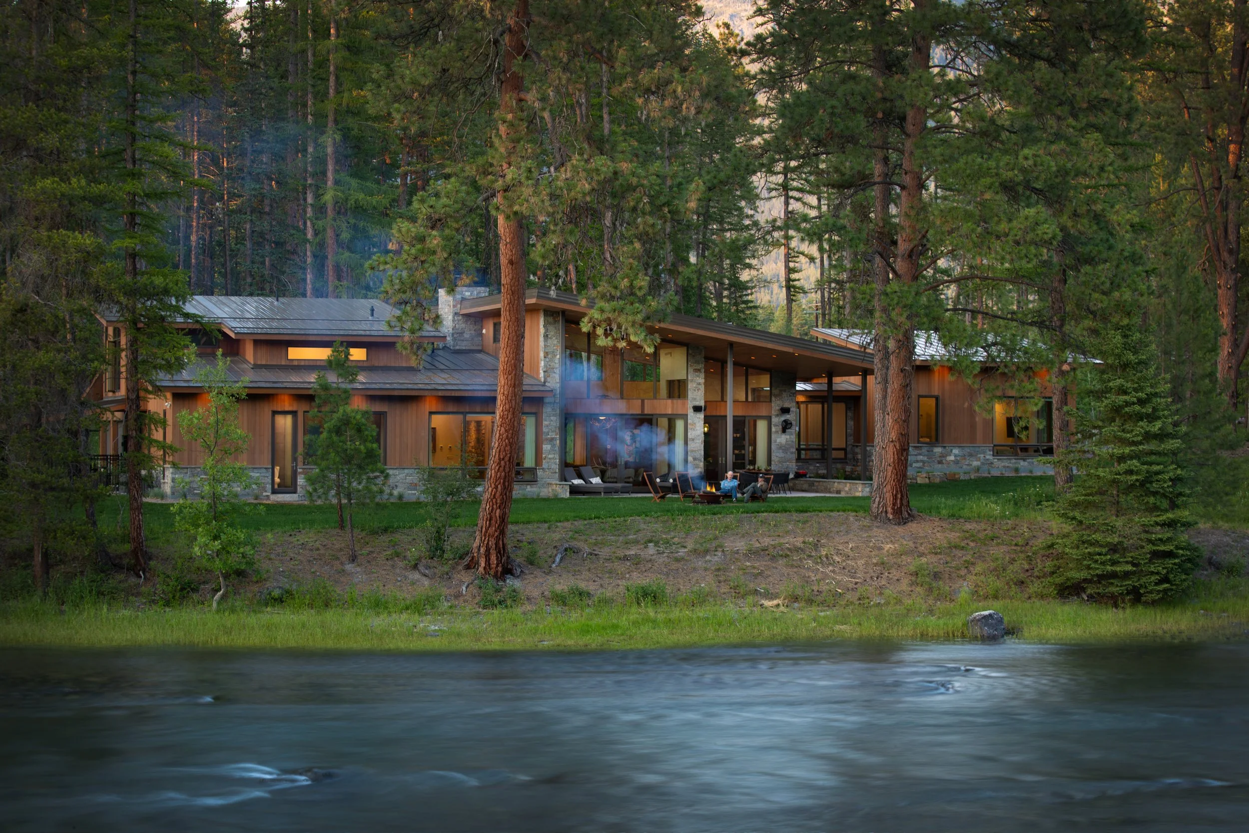 A modern house with wooden exterior and large windows, situated among tall pine trees along a river, with people sitting around a fire outside. Exterior stonework completed by Lindberg Masonry in Kalispell Montana.
