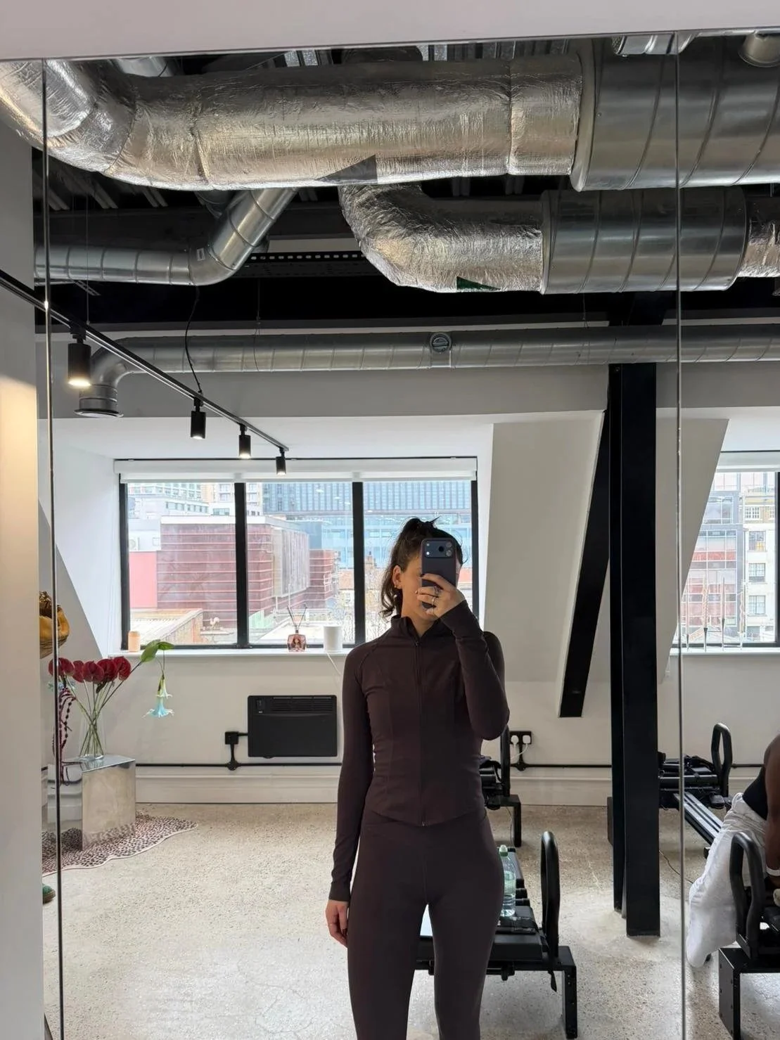 A woman taking a mirror selfie in a modern gym with exposed ductwork ceiling, large windows with cityscape view, and exercise equipment in the background.