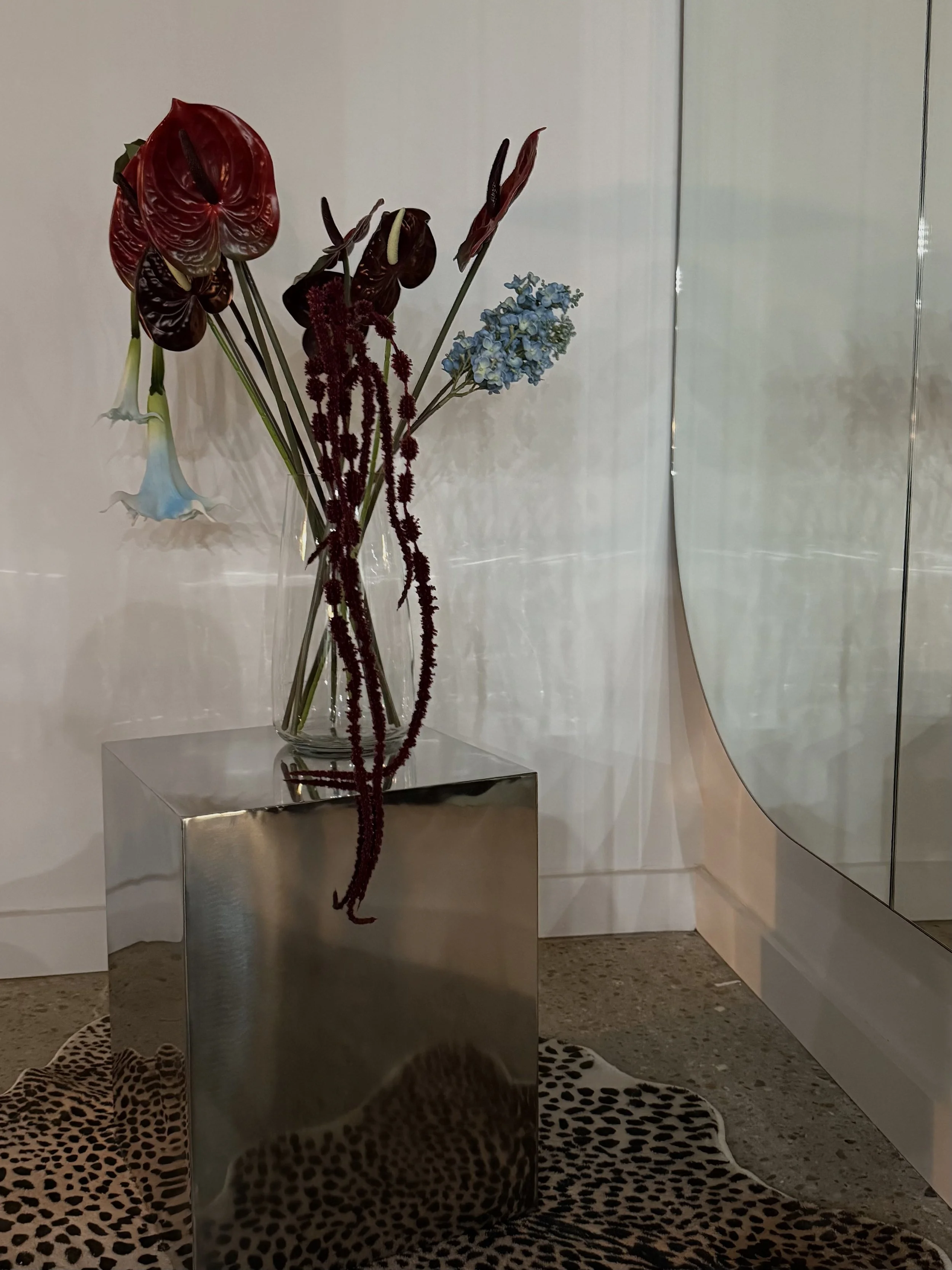 A glass vase with various flowers, including red anthuriums, a blue hydrangea, and dark red amaranth, sits on a reflective silver cube. The setting is indoors with white walls, a mirror, and a leopard print rug.