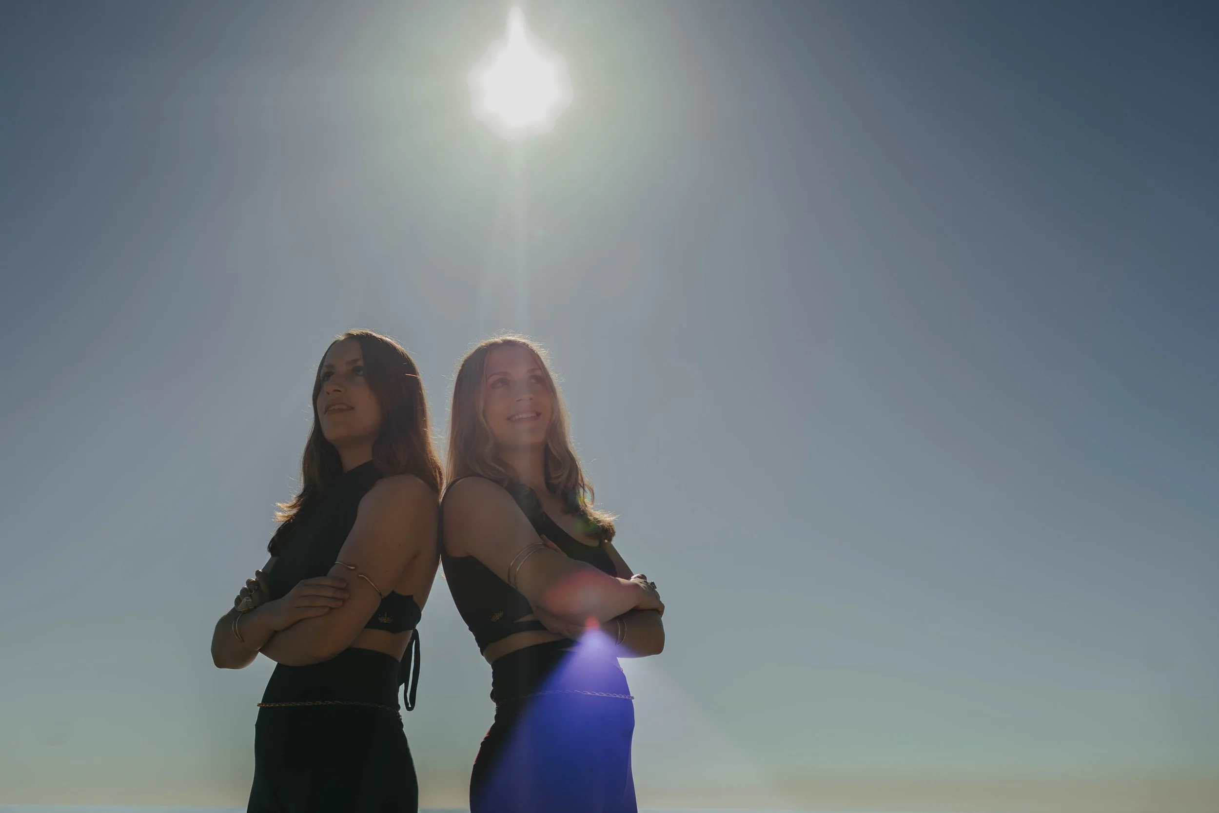 Marley and Lacey stand back to back with their arms crossed. The Sun is beaming above them depicting a portal.