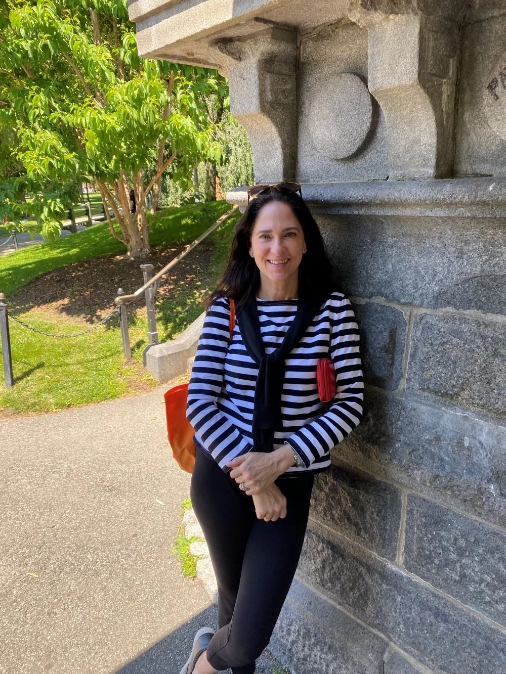 A woman with black hair in a striped shirt and black pants smiling while leaning against a stone wall outdoors on a sunny day.
